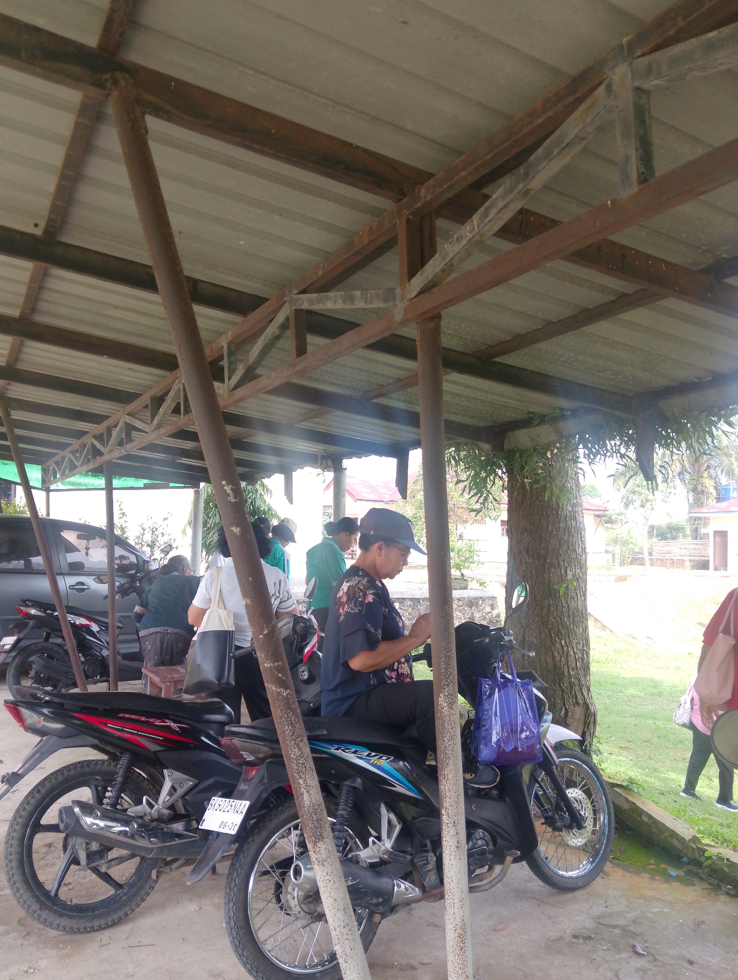 The image depicts an outdoor scene under a covered structure, likely a parking area or shelter, in Serbelawan, Indonesia. The structure is supported by rusty metal poles and beams, with a corrugated metal roof. Several motorcycles are parked within the shelter, including two in the foreground. One is a black motorcycle with red accents, identified by the license plate "BK 6025 V4A". A person wearing a dark blue shirt with a floral pattern and a black cap is seated on this motorcycle, looking down at their phone. A purple plastic bag is hanging from the handlebars.
In the background, a silver car is parked next to a motorcycle. Several other individuals are visible, some standing and some appearing to be in conversation. A person in a white shirt is holding a white tote bag, and another individual is wearing a green shirt. The environment beyond the shelter includes trees, grass, and buildings in the distance, suggesting a semi-urban or rural setting. The lighting indicates it is daytime, with some dappled sunlight filtering through the structure.