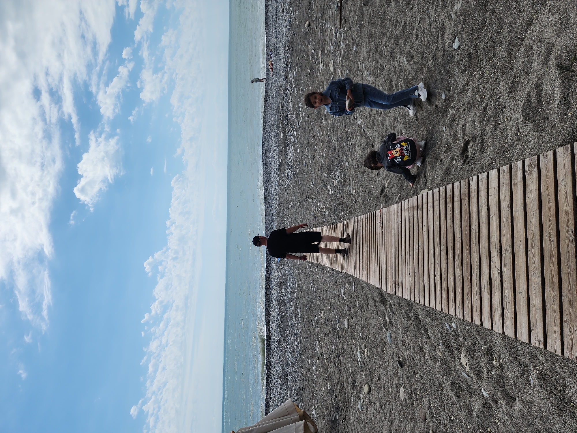 This image captures a lively beach scene under a partly cloudy sky, with a wooden walkway leading towards the calm sea. The sky, a bright blue, is dotted with various white clouds, indicating fair weather. The light blue-green sea extends horizontally across the middle of the frame, meeting a dark pebble and sand beach.
A prominent feature in the foreground is a wide wooden walkway, constructed from parallel planks, that stretches from the bottom right corner of the frame towards the sea. Three individuals are central to the scene. A man in a black t-shirt, shorts, and a dark cap is seen walking on this wooden path, moving away from the viewer towards the water.
On the sandy part of the beach, closer to the sea, a woman dressed in a denim jacket and jeans stands, facing the camera with a slight smile and holding a small, light-colored object in her hand, possibly a shell or a pebble. Adjacent to the wooden walkway, a child is kneeling or crouching on the dark sand. The child wears a black jacket with a colorful cartoon character design, resembling Mickey Mouse, on the back, and a pink skirt. They appear to be engaged in playing or examining something on the ground. In the distance, near the water's edge, a few more faint figures can be discerned.
A portion of a beige beach umbrella or canopy is visible in the bottom left corner, hinting at beach amenities. The beach surface is a mix of dark sand and small, smooth pebbles. The overall atmosphere suggests a leisurely day at the beach, likely during daylight hours. No discernible text is visible other than the cartoon character design on the child's jacket. This scene is set on a beach in Sochi, Russia.