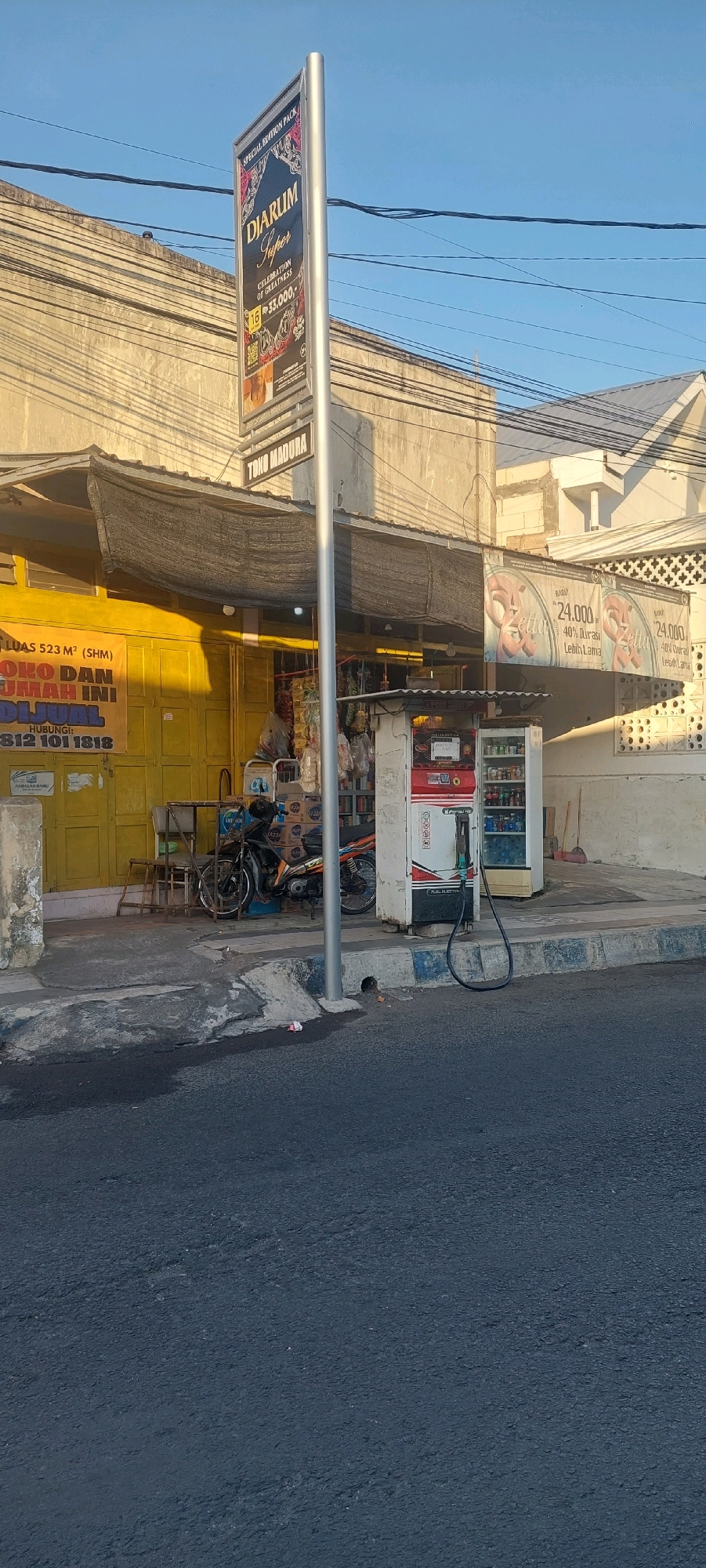 A street scene in Lumajang, Indonesia, captures the facade of a small shop with a yellow painted exterior. Above the shop, a black mesh awning provides shade. A prominent vertical sign displaying "DIARLUM Super" with ornate graphics and "Rp 33,000,-" is mounted on a silver pole in front of the building. A horizontal sign below it reads "TOKO MADURA".
In front of the shop, a small, white and red gasoline pump with a black hose is situated on the sidewalk. To the left of the pump, a motorcycle is parked. Various items, including bottled drinks and bags of goods, are visible inside a refrigerated display case next to the gas pump and through the shop entrance. A yellow banner on the shop front indicates "LUAS 523 M² (SHM)" and "TOKO DAN RUMAH INI DI JUAL" (Shop and House For Sale) with a contact number. The street in the foreground is paved with asphalt. The sky above is clear and blue.
