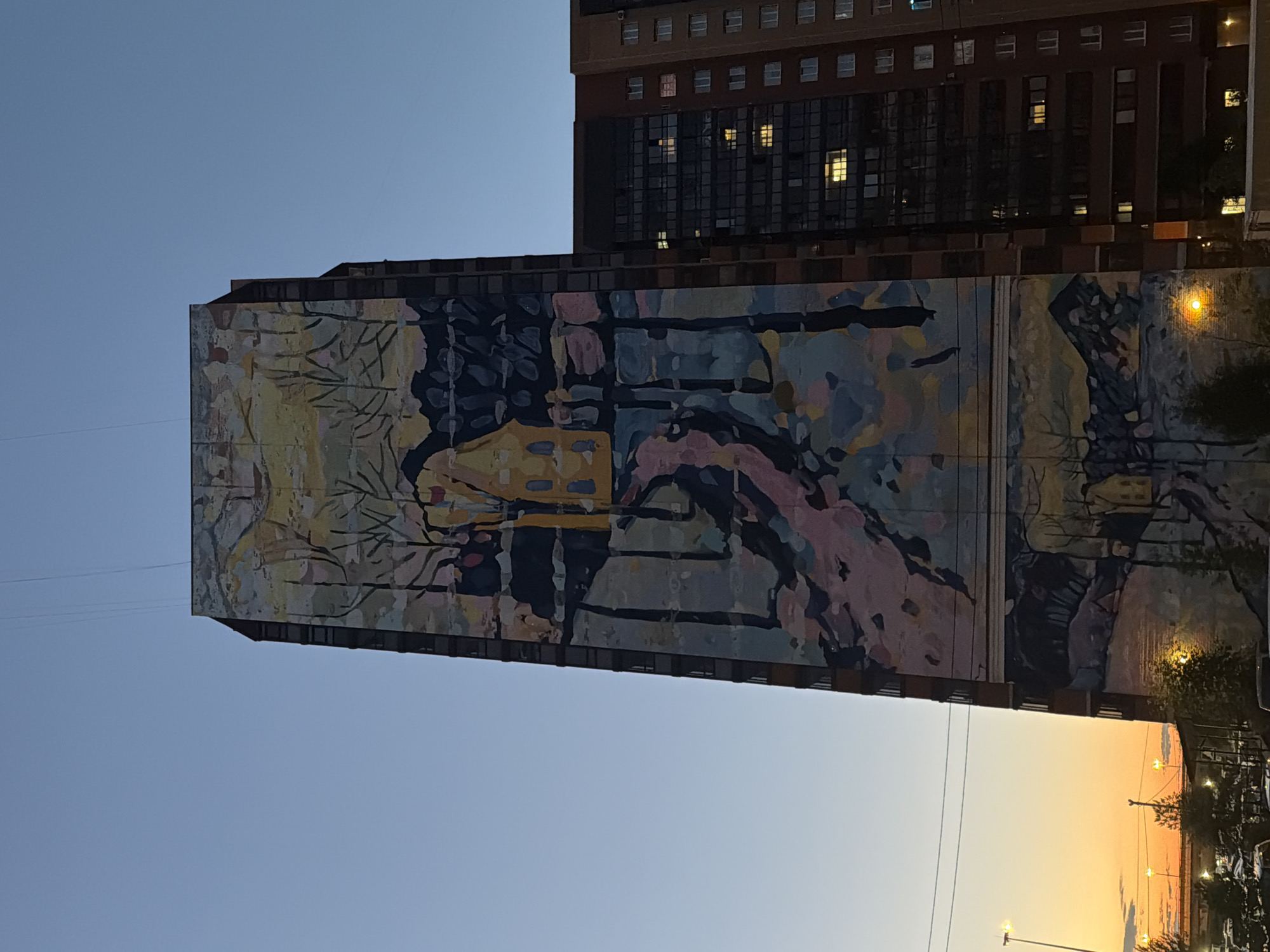 An extreme low-angle, full shot captures a mural painted on the side of a tall, slender, multi-story building at dusk. The mural, rendered in an impressionistic style with a palette of muted colors including yellow, pink, blue, and black, depicts a landscape with trees, a path, and a small house. The mural covers a significant portion of the building's facade. To the right of the mural-covered building is a darker, more modern building with many windows, some of which are illuminated. The sky above is a gradient of light blue to pale orange near the horizon, indicating sunset. In the foreground, the tops of trees and streetlights are visible, along with the silhouette of parked cars. Utility wires are visible stretching across the upper portion of the sky.