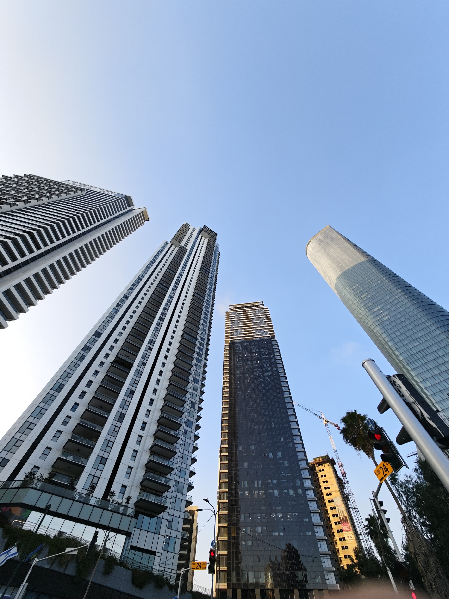 This is a low-angle, wide-shot photograph taken in the city of Givatayim, Israel, depicting a cluster of modern skyscrapers against a clear blue sky. The buildings are tall and predominantly feature glass and white facades.
In the foreground and left of the frame, a long, thin skyscraper with repeating balconies and dark windows is angled upwards. To its right, a wider skyscraper with a more textured white facade and numerous balconies is also visible, extending further into the frame.
Further to the right, a dark, slender skyscraper with a reflective glass facade rises steeply. Next to it, a distinctive, rounded cylindrical skyscraper with a light blue, glass-covered exterior dominates the right side of the image. Its upper section appears to have a segmented or paneled design, with a lighter color than the lower part.
In the mid-ground, a light-colored building under construction is visible, with a crane towering over it. Palm trees and other foliage are scattered around the base of the skyscrapers, suggesting an urban park or landscaped area.
Traffic lights and street signs are present in the lower right corner, with one sign displaying "24" in yellow. The overall scene conveys a sense of modern urban development and architecture.