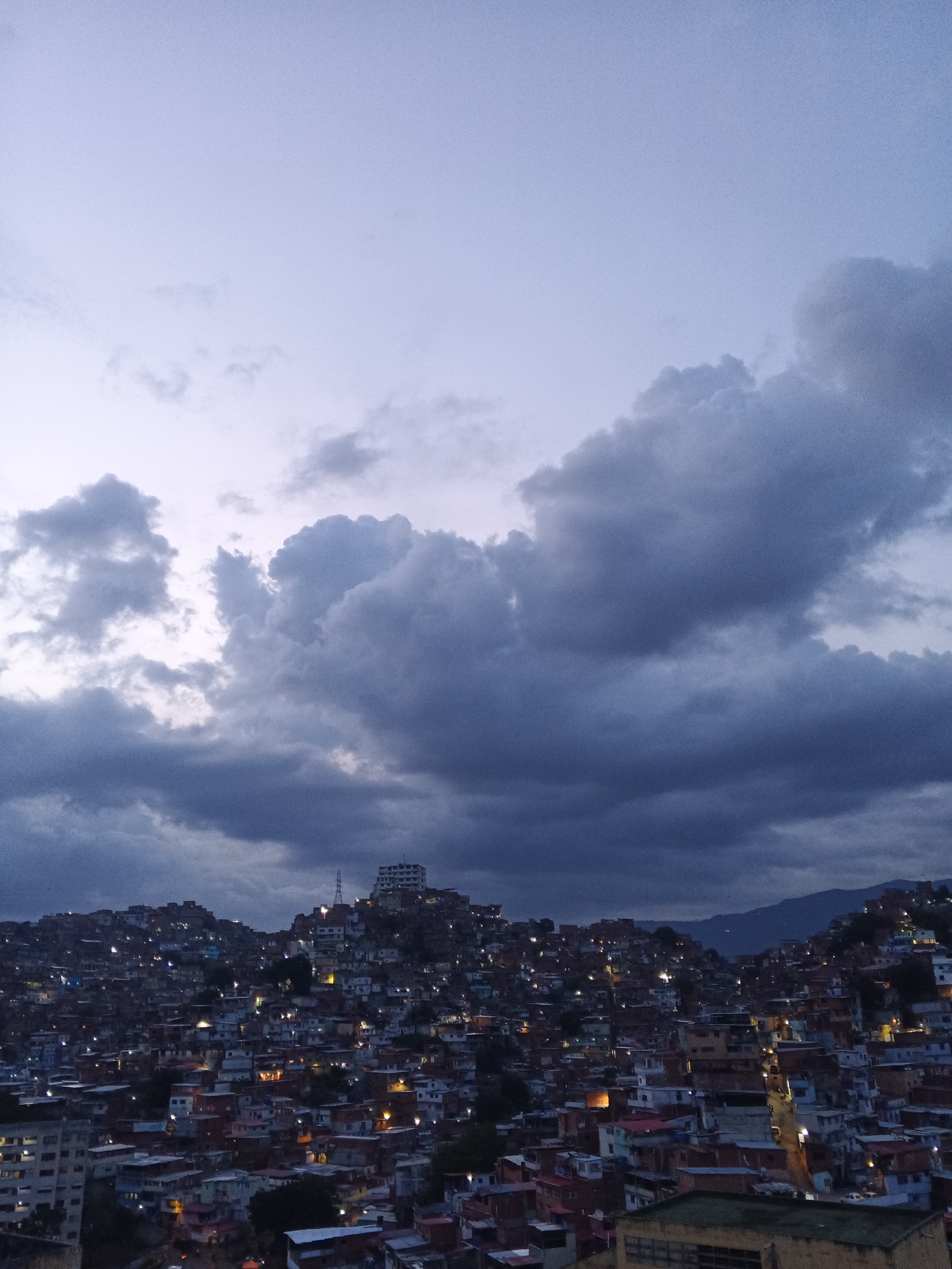 The image shows a densely populated urban area in Caracas, Venezuela, during the early evening or twilight hours. The foreground and midground consist of closely packed buildings of varying sizes and colors, likely residential structures. Numerous lights emanate from within these buildings, suggesting activity and inhabitation. The structures are built on uneven terrain, characteristic of the hills surrounding Caracas. In the background, more buildings climb the hills, culminating in a prominent multi-story building with a tower on a high point. Dark, heavy clouds dominate the sky, giving the scene a somber or dramatic atmosphere. In the far background, mountains are visible, suggesting the valley location of the city. There are no people visibly in the image.