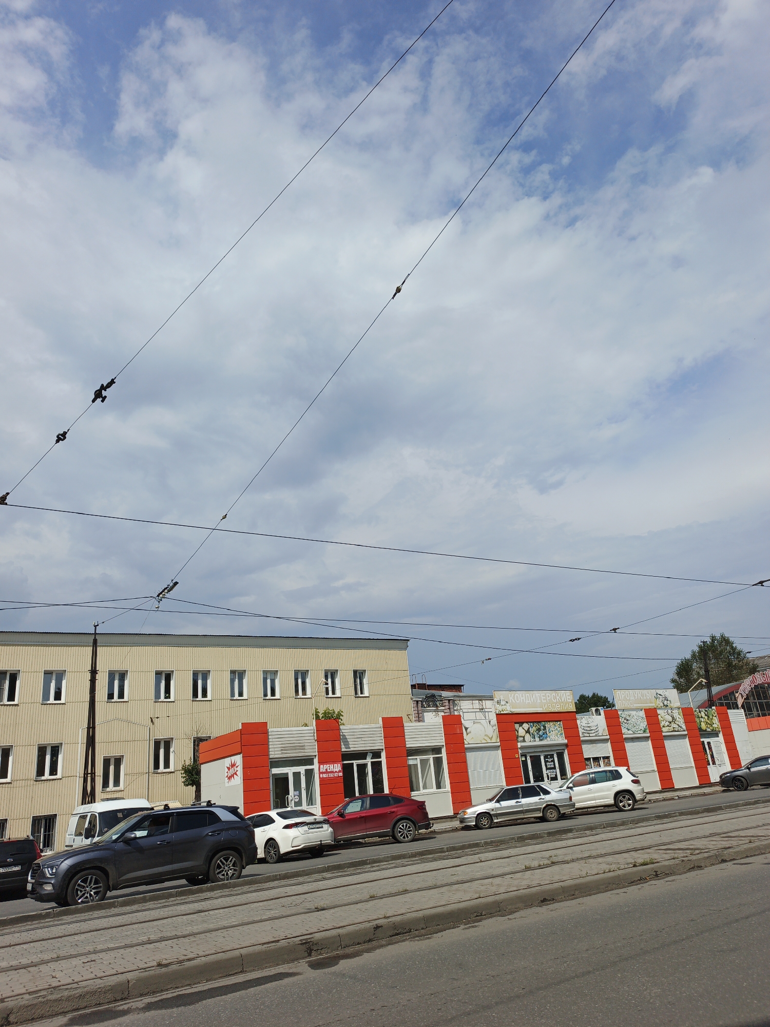 A street-level, medium-shot, eye-level, outdoor, daytime, vertical, full-color, slightly warped, wide-angle photograph shows a view along a street in Zlatoust, Russia, with a line of parked cars and storefronts. Overhead, multiple power lines crisscross the sky, which is mostly blue with scattered white clouds. The buildings are primarily two stories. The building on the left is a light yellow, long structure with many evenly spaced white-framed windows on the upper floor. Below the windows are what appear to be trolley tracks and a sidewalk. In front of this building, parked vehicles are visible. A dark gray SUV is in the foreground on the left. To its right is a white van, followed by a silver sedan, a red sedan, and a white SUV. A second building, to the right of the first, has a modern design with a red and white striped facade. This building has several large windows or glass doors, and signage is visible above some of the storefronts. One sign says "КОНДИТЕРСКИЕ ИЗДЕЛИЯ" (Confectionery Products), and another reads "ПРОДУКТЫ ПЛЮС" (Products Plus). A dark gray sedan is parked in front of this building on the right. The street surface consists of asphalt and what appears to be a tram track or trolley line. The overall impression is a typical urban street scene on a bright, cloudy day.