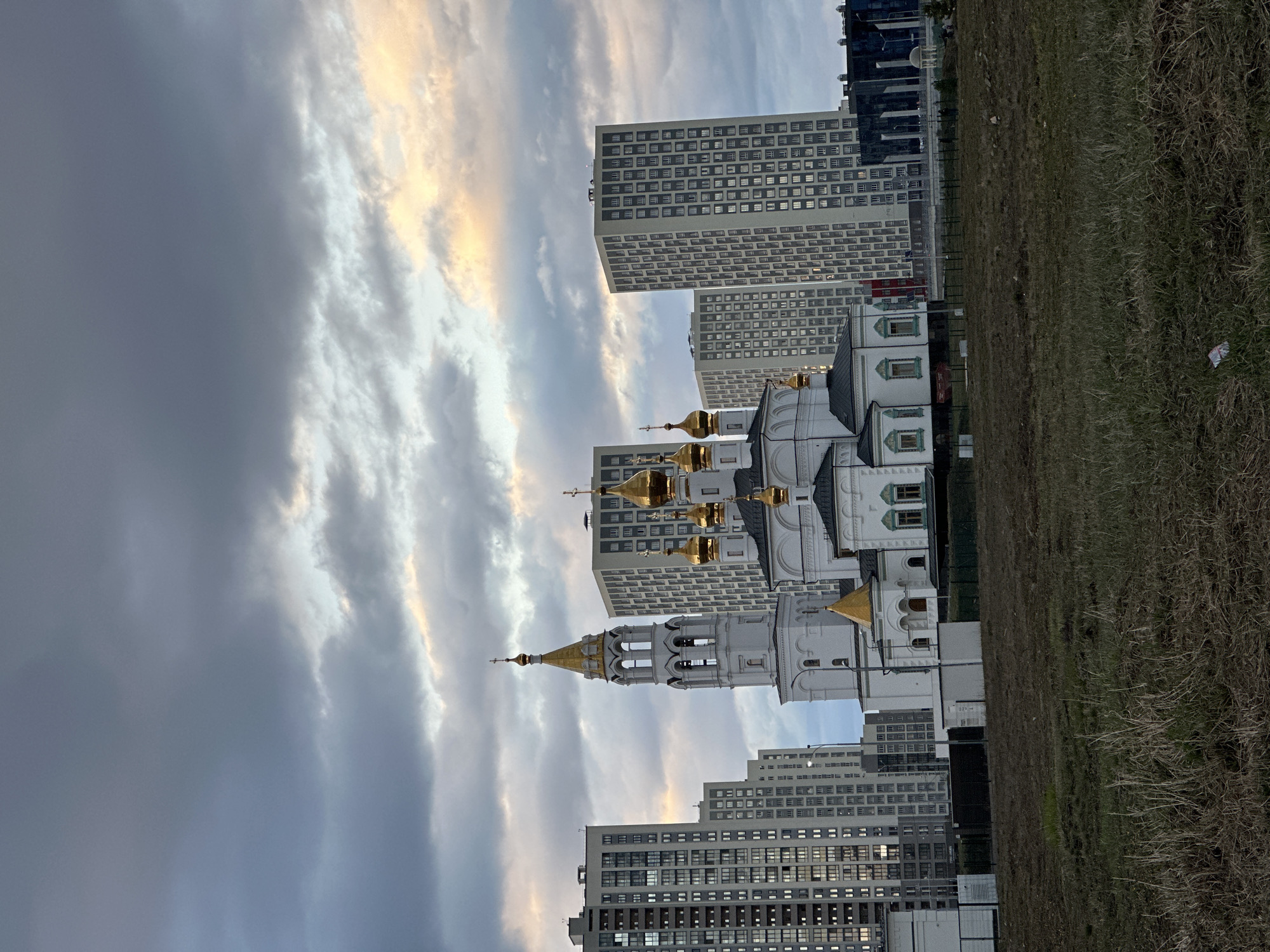 The image shows a view of the Church on Blood in Yekaterinburg, Russia, framed against the backdrop of modern high-rise buildings and a cloudy sky. The church's white walls and gold domes contrast with the gray, rectangular architecture of the surrounding buildings. A patch of dry, brown grass occupies the foreground on the right side of the frame. The sky is filled with striated clouds, suggesting either early morning or late evening light. The photograph appears to be taken at ground level.