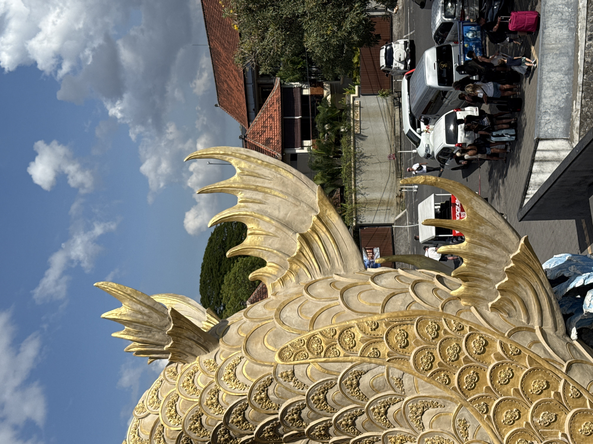 The image captures a scene near the Bali Ngurah Rai International Airport in Denpasar, Indonesia. In the foreground is a large, golden fish statue with detailed scales and decorative patterns, occupying a significant portion of the frame.
In the background, a street scene unfolds. There are several white vans parked along the roadside, with groups of people standing beside and near the vehicles. Some individuals are loading or unloading luggage, including a bright pink suitcase, suggesting they are likely tourists arriving or departing. The background also includes a building with a red-tiled roof, greenery and palm trees and a clear sky with scattered clouds.