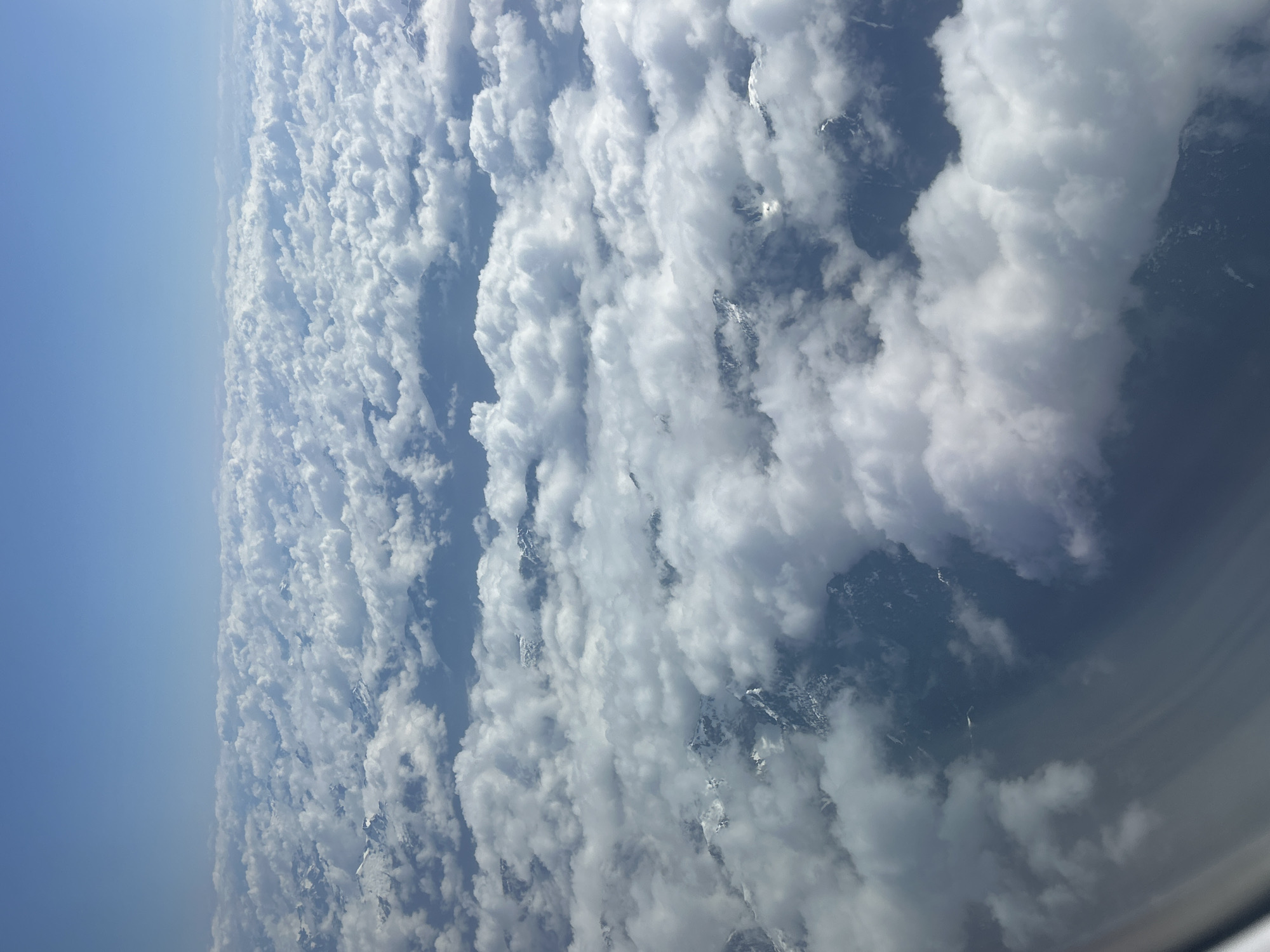 The image presents an aerial view of clouds, seemingly taken from an airplane window. The left side of the image shows a clear blue sky transitioning into a dense layer of white, puffy clouds. Below the clouds, hints of snow-capped mountains can be discerned, suggesting a high-altitude perspective. There is no evidence or indication in this image of anything in Kelowna, Canada.