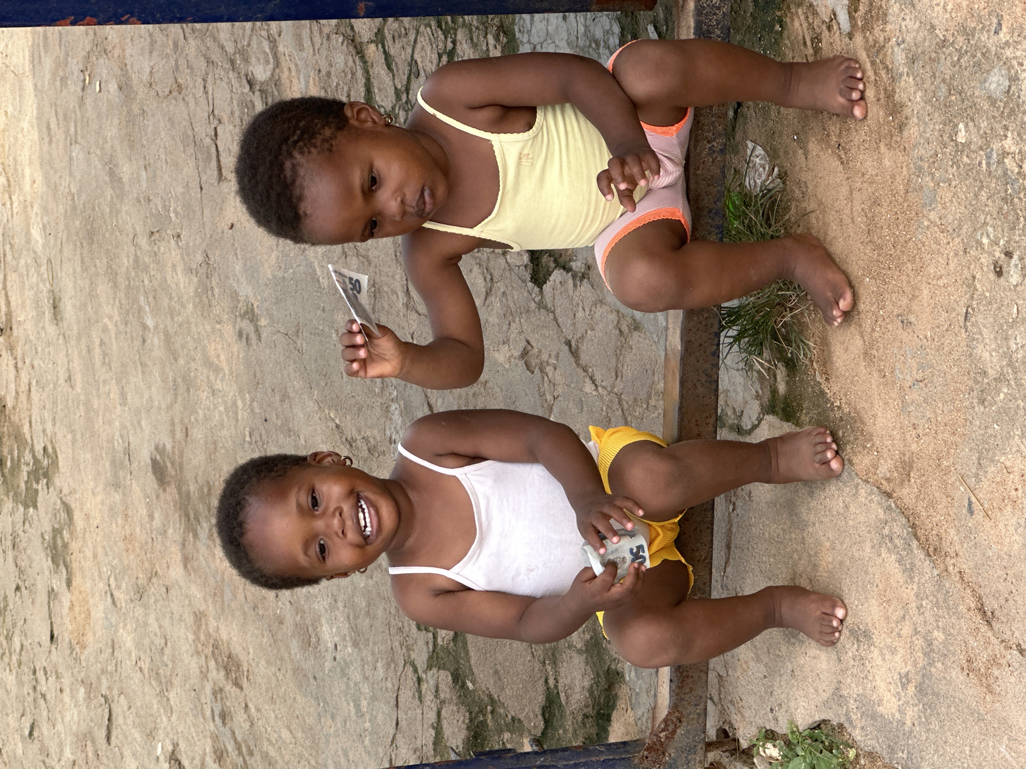 Two young girls are sitting on a concrete surface outside. The girl in the foreground is wearing a white tank top and yellow shorts. She is smiling widely and holding a small piece of paper in her hands. The girl in the background is wearing a yellow tank top and pink and white shorts. She is looking down at a small piece of paper she is holding. Both girls have dark skin and dark hair. The concrete surface is rough and appears to be outdoors, with some patches of dirt and grass visible. The scene is lit by natural light, suggesting it is daytime. The girls appear happy and engaged in their activity. The visible text on the paper held by the girl in the foreground appears to be "50". The setting appears to be in Narayi, Nigeria, with the presence of a blue metal railing in the background, common in such areas.