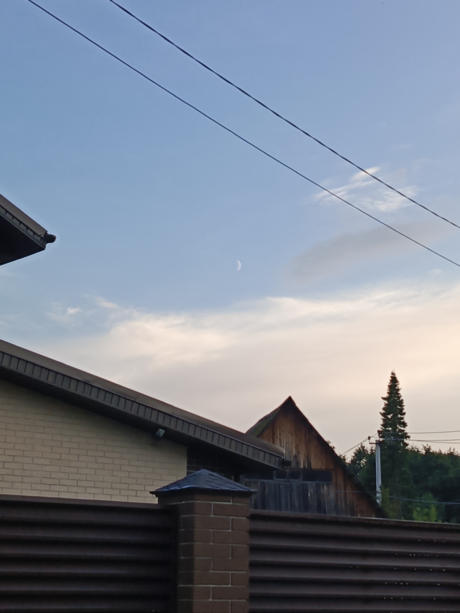 A low-angle shot captures a crescent moon visible in a pale blue sky with scattered clouds, above a suburban landscape in Chaikovsky, Russia. Two power lines traverse the sky diagonally. In the foreground, the dark, corrugated metal fence of a property runs horizontally across the image. Above the fence, the light-colored brick wall and dark-shingled roof of a building are visible on the left. To the right, a dark wooden-shingled roof with a triangular gable is seen, behind which a tall, dark green evergreen tree stands. Behind the tree, a utility pole with wires is visible amidst a dense line of green trees. The scene appears to be set at dusk or dawn, given the sky's coloration and the moon's visibility.