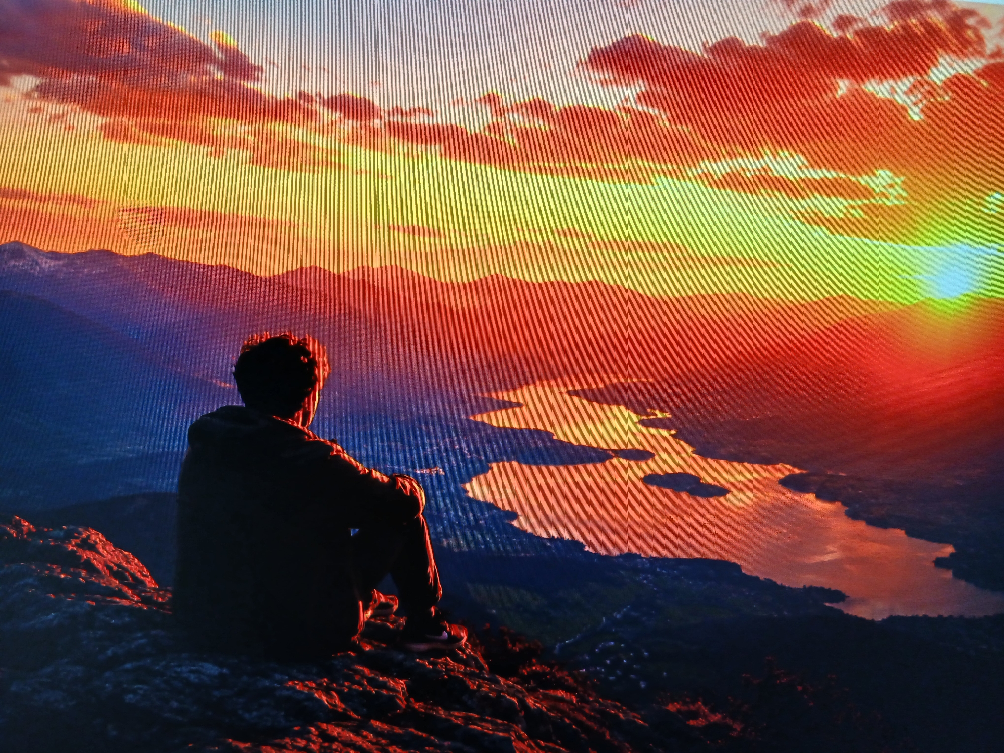 The image depicts a breathtaking sunset over a mountainous landscape with a body of water winding through it. In the foreground, a lone individual sits on a rocky outcrop, silhouetted against the vibrant sky. The sky is a spectacle of fiery oranges, yellows, and reds, with dramatic clouds illuminated by the setting sun. The mountains in the distance are bathed in shades of purple and blue, creating a sense of depth and grandeur. The water reflects the colors of the sunset, appearing as a ribbon of light cutting through the darker terrain. The scene evokes a sense of peace and contemplation. There is no information provided in the image or its crops that would allow for identification of the city of Pleven, Bulgaria.