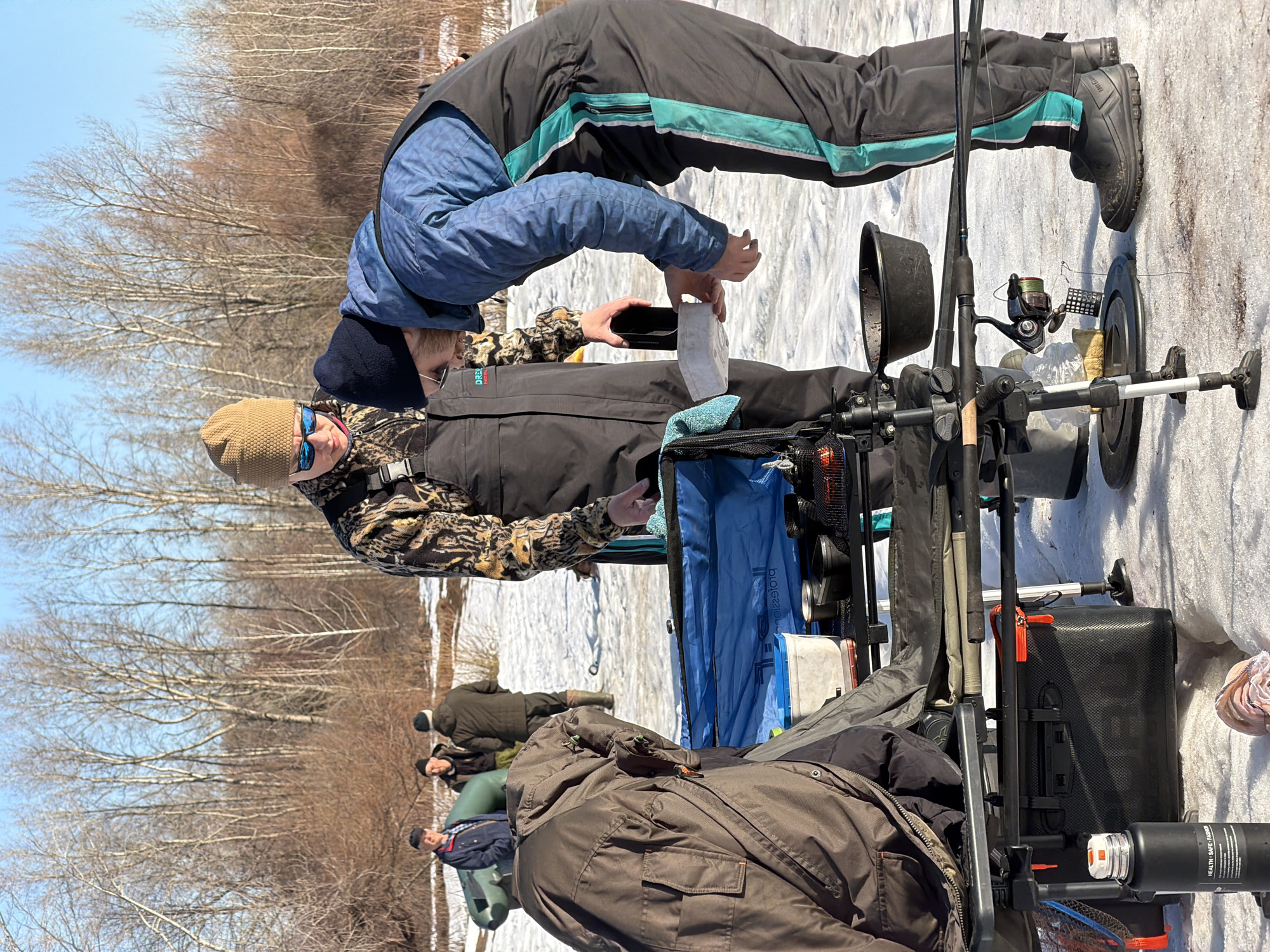 The image depicts a winter scene with several people engaged in ice fishing. Two individuals are the focus: one is leaning over towards a fishing setup, possibly preparing bait or tackle, while the other, wearing a hat and sunglasses, appears to be overseeing the activity. The fishing equipment includes rods, reels, and a tackle box mounted on a two-wheeled cart. Other individuals are visible in the background, also participating in ice fishing. The environment is snowy, with bare trees indicating winter. The scene is likely outdoors, possibly on a frozen body of water. The city of Chaikovsky, Russia is mentioned as the location.