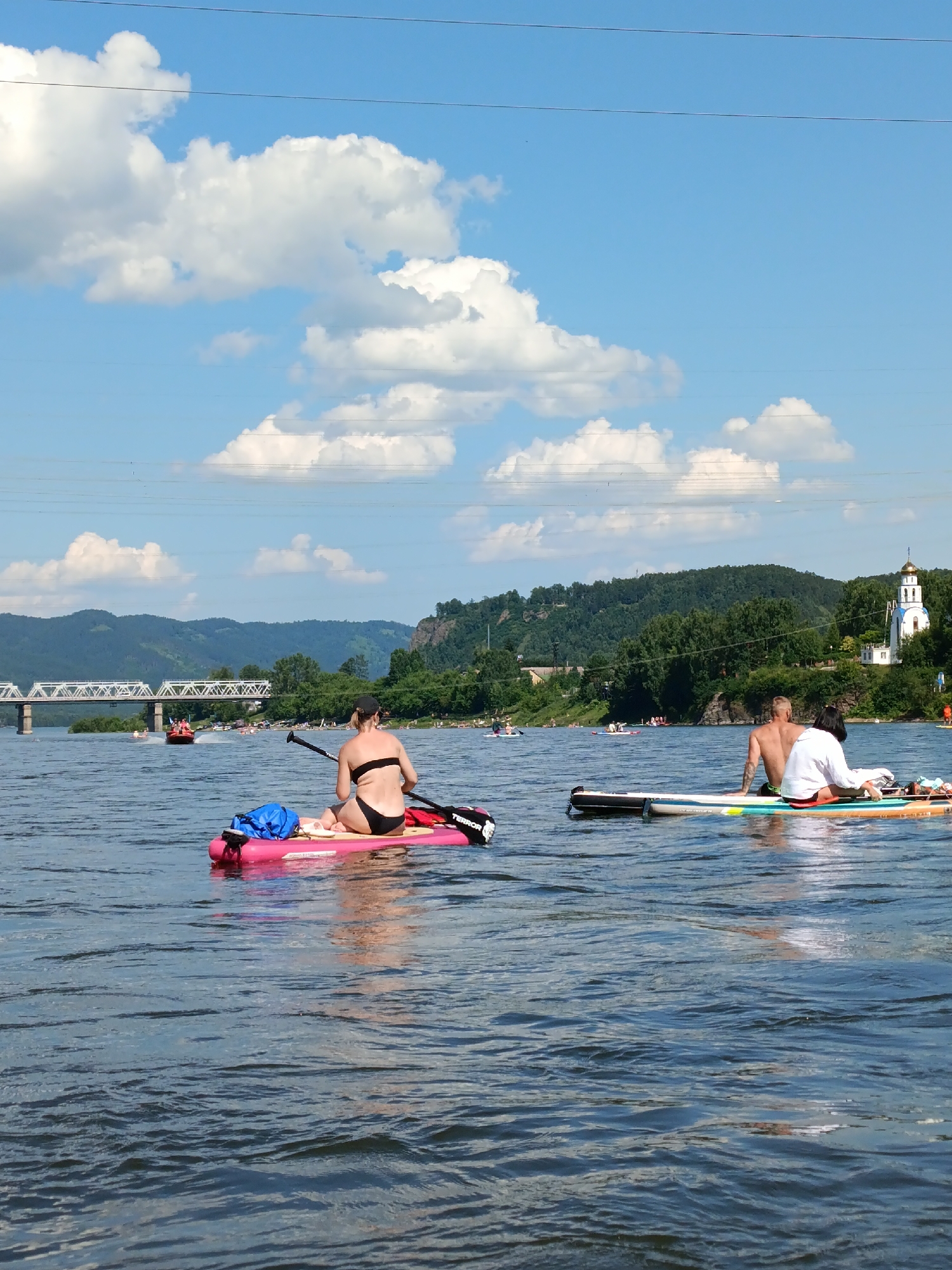 In this image, taken at Ust-Mana, Russia, a bright, sunny day is depicted on a body of water, likely a river, with a blue sky dotted with fluffy white clouds. The water's surface is rippled and reflects the light. In the foreground, two people are on separate paddleboards. On the left, a person in a black bikini is seated on a pink paddleboard, facing away from the camera, with a blue bag and a paddle beside them. On the right, a couple is seated on a paddleboard, facing the water. The man is shirtless, and the woman is wearing a white shirt. In the mid-ground, several other paddleboarders and a small boat are visible on the water. In the background, a lush, green forested hillside rises, and a white building with a golden dome, resembling a church, stands on the hill. A large white bridge with multiple spans crosses the water in the distance, and power lines stretch across the sky. The overall scene suggests a recreational day by the water.