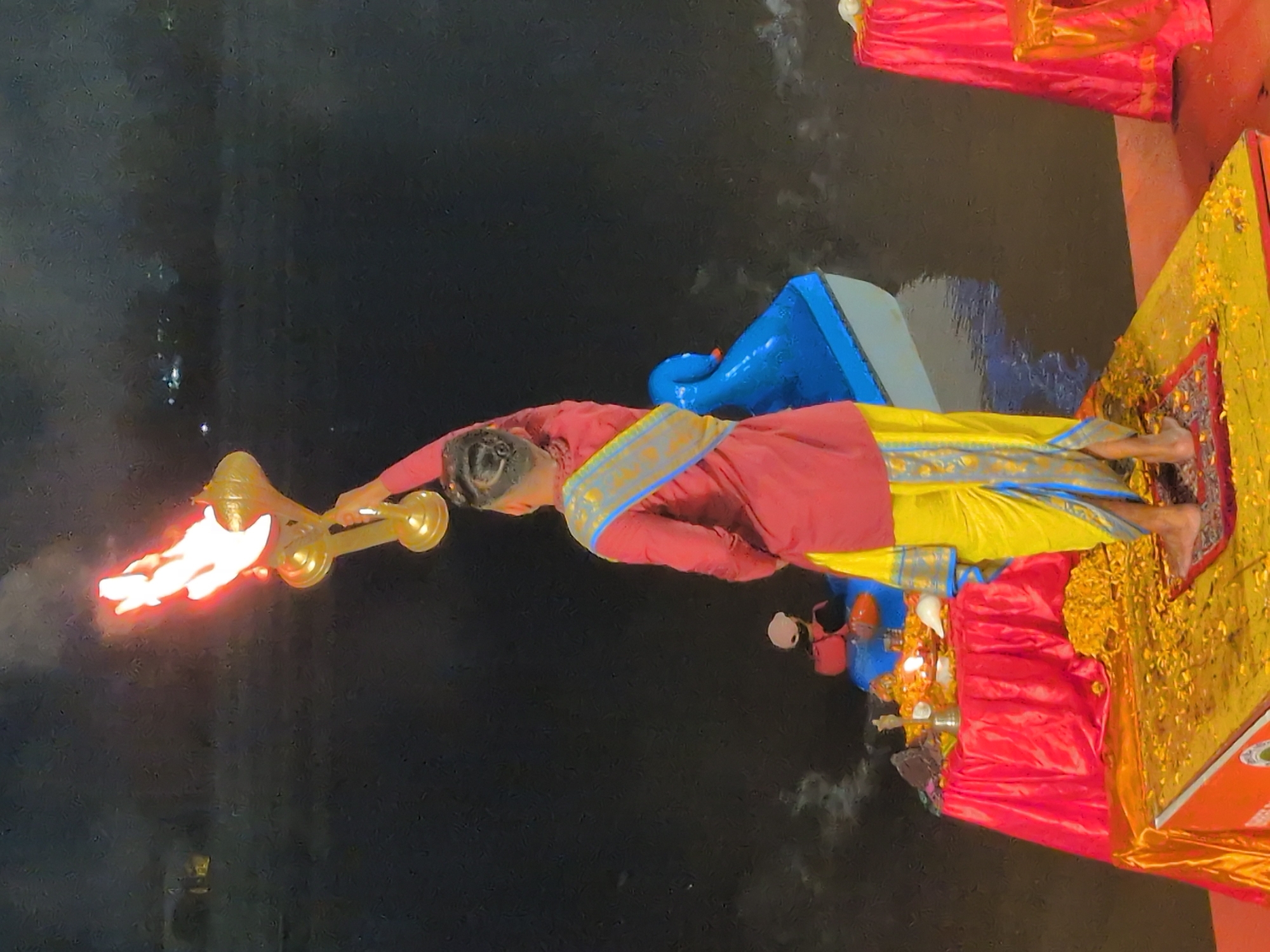 A rear view of a person in traditional Indian attire, including a maroon shirt and a yellow dhoti with blue trim, stands on a platform covered in yellow petals. The person holds a golden ceremonial lamp with flames aloft. The platform is situated near a body of water, possibly the Tawi River in Jammu, India, where a blue boat with a decorative swan head is visible. In the background, lights twinkle, suggesting the scene occurs at night or dusk. To the left, a red table is draped with a red cloth and adorned with flowers and religious items, including a conch shell and a lit oil lamp. A red cloth-covered table is also visible on the right. The overall atmosphere suggests a religious ceremony or ritual.