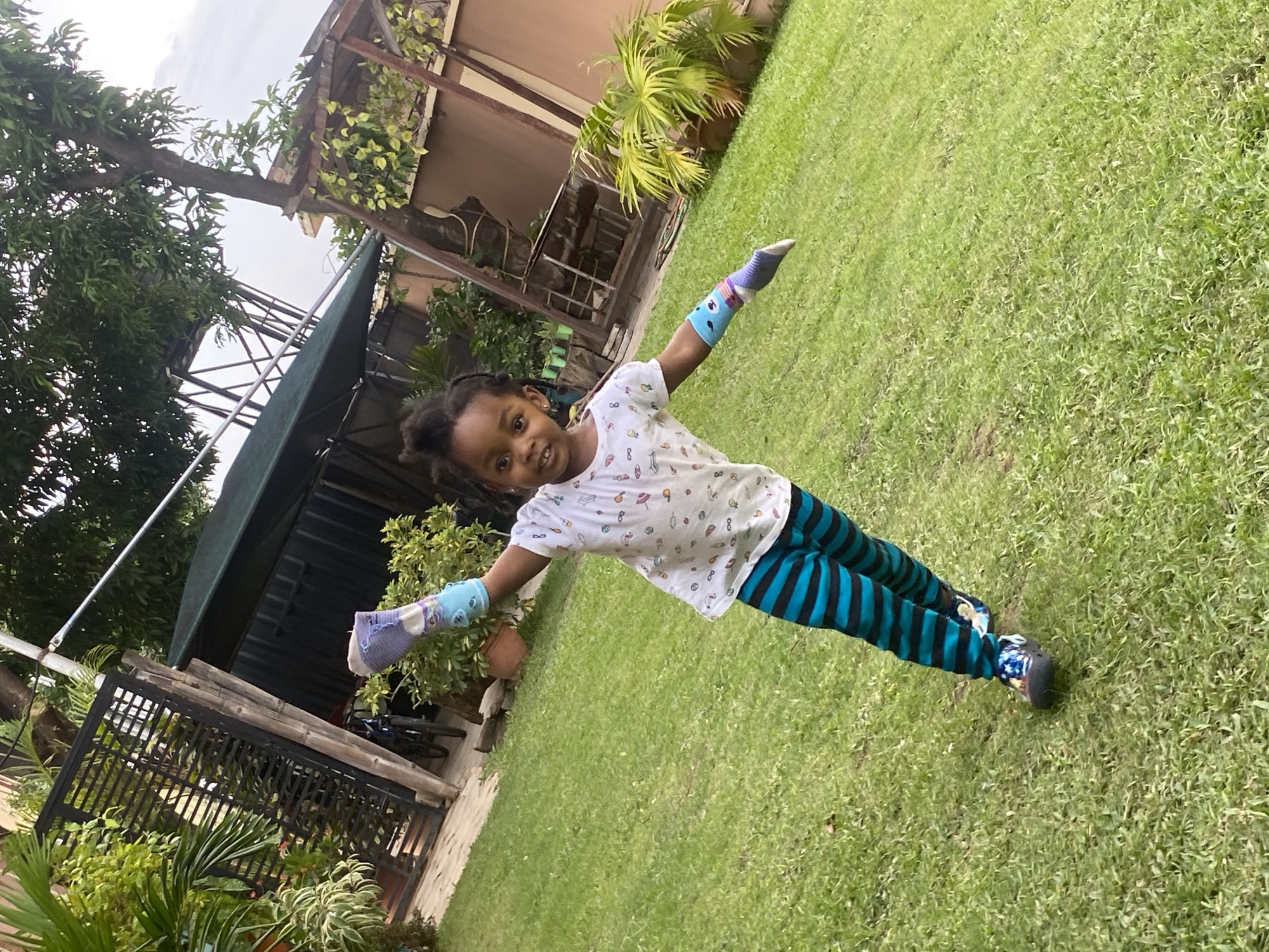A young girl stands on a green lawn in what appears to be an outdoor space. She is wearing a white t-shirt with a pattern and blue and black striped pants. She has socks on her arms. There is a building with a black metal fence, trees, and other greenery in the background. The location is assumed to be Barden Yana, Nigeria.