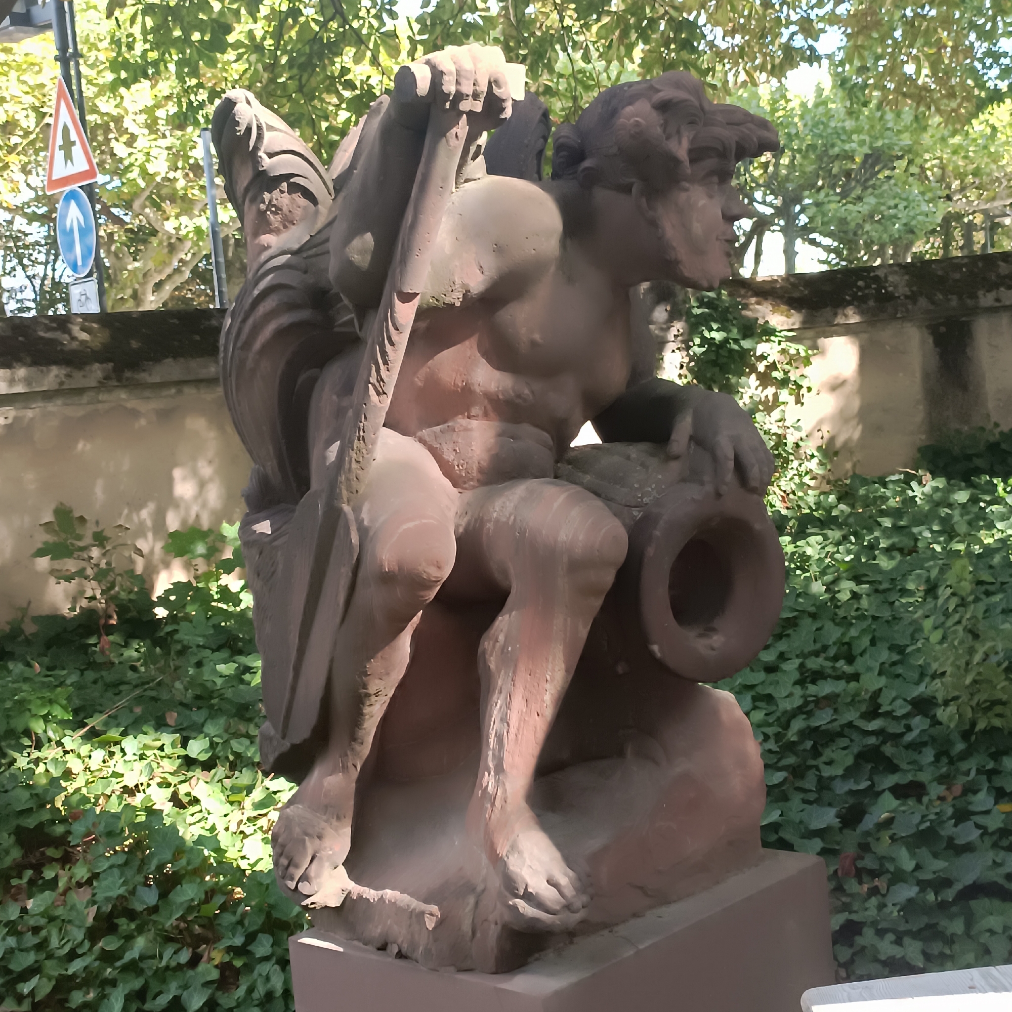 The image shows a stone sculpture of a winged figure with a large amphora. The sculpture is situated outdoors, with lush green ivy covering the ground and a stone wall in the background. Trees with green foliage are visible above the wall. Two traffic signs are present to the left of the sculpture; one is a red and white triangle with a bird symbol, and the other is a blue circular sign with a white arrow pointing upwards. The sculpture appears to be made of reddish-brown stone and is partially weathered. The figure's musculature is well-defined, and its wings are large and detailed. The amphora is large and round with a narrow neck. Sunlight casts shadows on the sculpture and the surrounding vegetation, indicating it is daytime. The overall impression is of a classical-style sculpture in a park-like setting. This sculpture is located in the Grüneburgpark in Frankfurt, Germany.