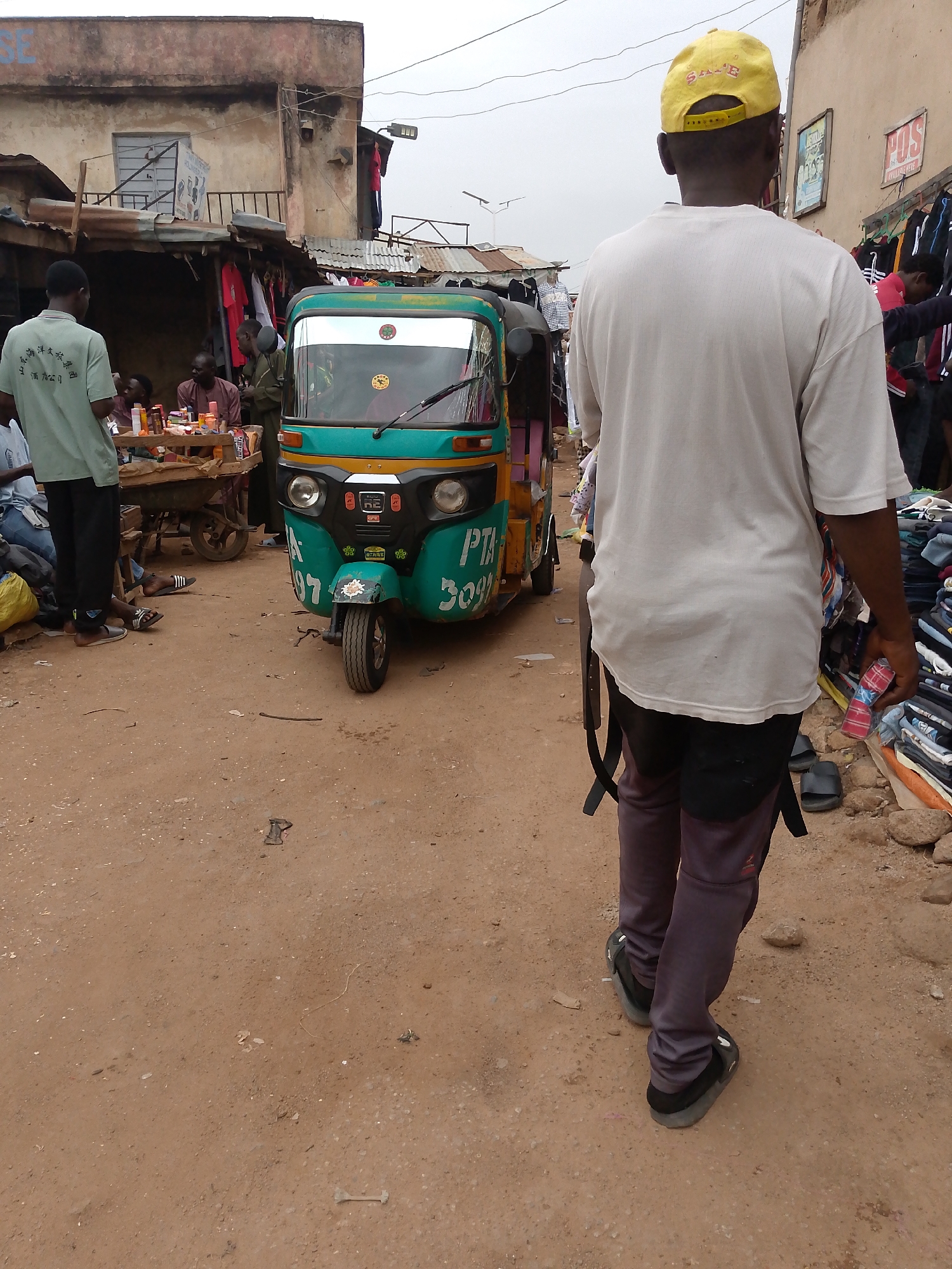 The image depicts a busy scene on a dirt road in Bukuru, Nigeria. A green and orange three-wheeled vehicle, a "Keke Napep" or "auto-rickshaw," occupies the center of the frame. It is labeled "PTA 3097."
Several individuals are present. A man in a white t-shirt and yellow cap walks toward the viewer. To the left, another man in a green shirt stands by a makeshift table where others are seated, possibly eating or drinking. People are gathered around the vehicle and in nearby stalls.
The environment is a marketplace. Merchandise, primarily clothing, is displayed along the roadside. Buildings flank the scene, with a multi-story structure on the left and shops on the right. Overhead electrical wires are visible against a somewhat overcast sky. The ground is a reddish-brown dirt surface.