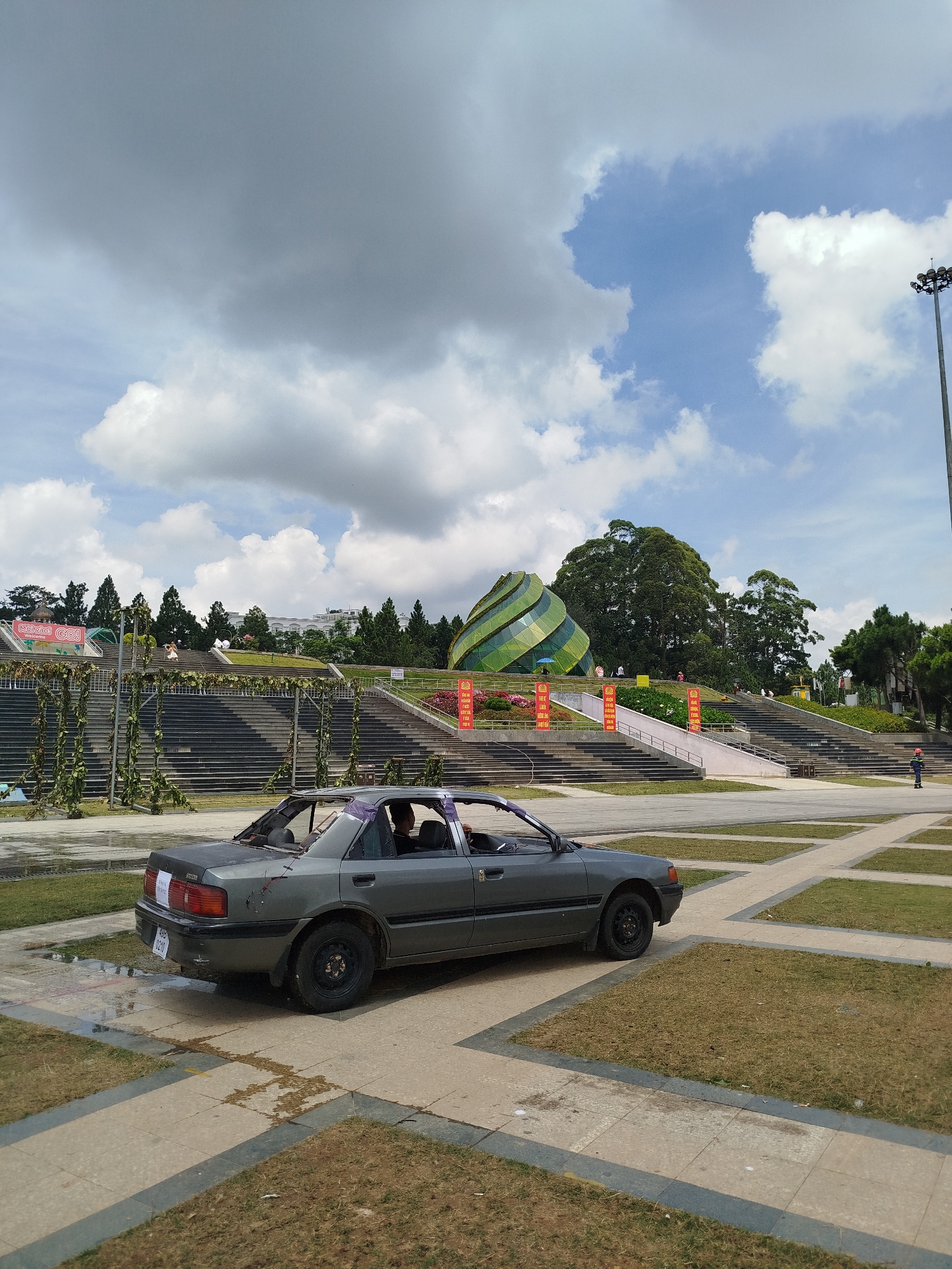 In Da Lat, Vietnam, a grey, dilapidated sedan with a damaged roof sits on a paved area adjacent to a large outdoor amphitheater. The car appears to have been spray-painted black on the trunk and possibly the roof, with visible damage to the rear window and a purple tape securing the edges. A white sticker with red writing is affixed to the rear bumper.
The amphitheater features tiered seating made of dark stone or concrete, with green vines climbing up metal structures. Banners with Vietnamese text and images are displayed along the amphitheater’s sides. In the background, a striking, geometrically-shaped building with green and silver stripes rises against a backdrop of lush green trees. The sky is partly cloudy with a mix of grey and white formations. The ground in front of the amphitheater is a combination of grass, pavement, and some wet patches. A few individuals are visible in the distance, some near the building and others on the amphitheater steps. A tall lamppost stands to the right of the frame.