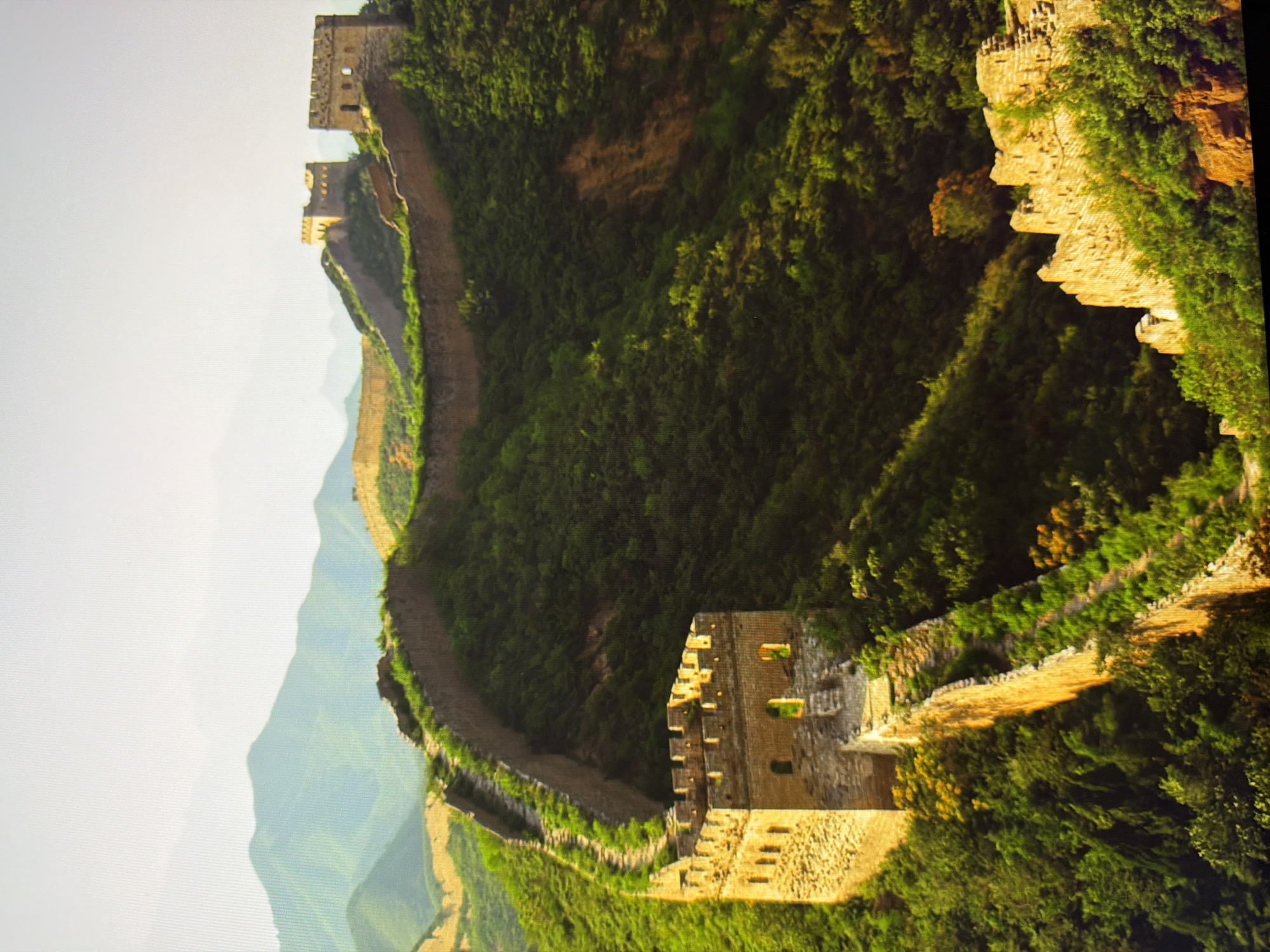 The image depicts the Great Wall of China, a historical fortification. The wall snakes across a lush, green mountainous landscape under a bright sky. Several watchtowers, constructed from weathered stone, punctuate the wall at intervals, some appearing partially ruined. The wall itself is made of stone and brick, with crenellated battlements visible. Vegetation, primarily trees and shrubs, covers the surrounding hillsides, with patches of exposed earth or rock also visible. The scene is bathed in warm light, suggesting it may be morning or late afternoon. There is no presence of people, animals, or any elements suggesting the city of Kyiv, Ukraine, which is geographically distant from this landmark.