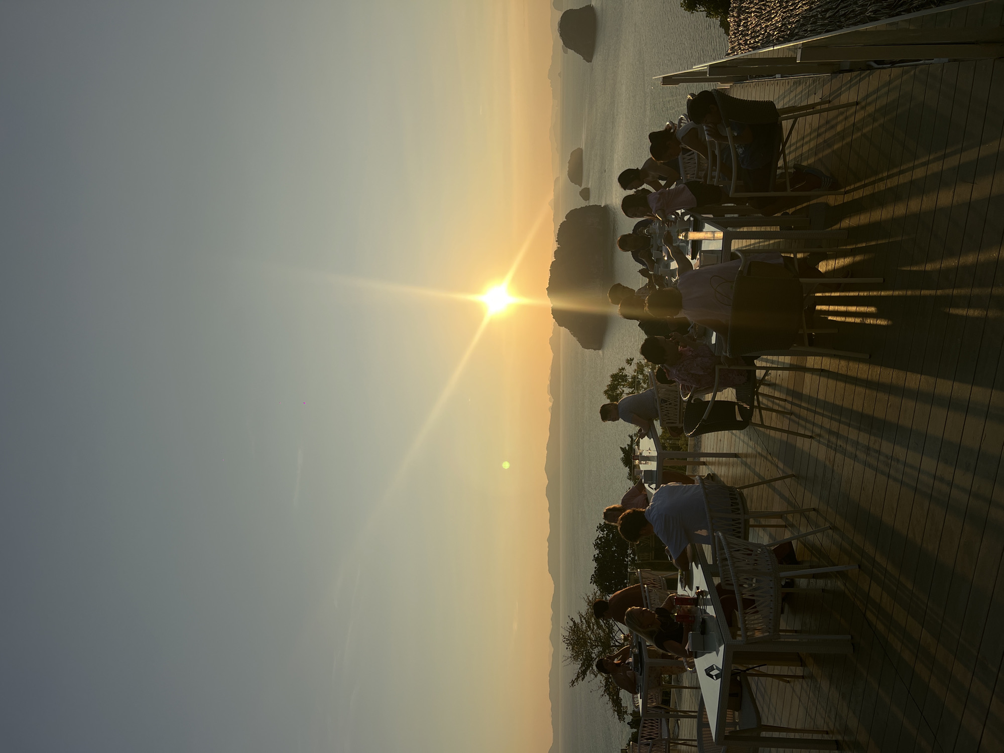 The image captures a sunset scene at a restaurant overlooking the sea. People are seated at tables, facing the sun. The restaurant is on a wooden deck. The sky is golden from the setting sun. In the water, there are several rock formations. The city is Khao Thong, Thailand.