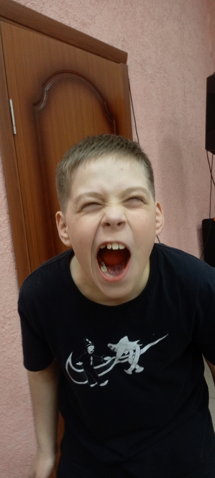 The scene is located within a building in Красный Маяк, Russia. A young male, appearing to be a child or pre-teen, is captured in a close-up, chest-up shot. He possesses short, light brown hair and is wearing a black short-sleeved t-shirt featuring a white graphic design depicting stylized figures. His eyes are forcefully closed, and his mouth is open wide, revealing his teeth and tongue, consistent with a loud vocalization such as a yell or scream. The immediate background to the left shows a section of a brown wooden door or cabinet with an arched decorative panel. The majority of the background consists of a textured, light reddish-pink wall. In the upper right, dark cables descend vertically, partially obscuring a dark, indistinct object, possibly a piece of electronic equipment.