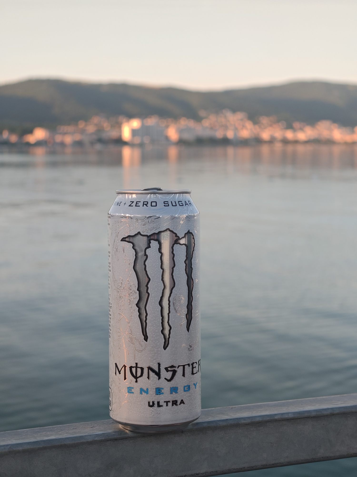 A can of Monster Energy Ultra drink rests on a gray metal railing. Behind the can is a body of water. Across the water is a city, possibly in Unknown, Unknown, which has a building with light reflecting off of it. Behind the city are the foothills of a mountain range. The sky is pale, hinting that the image may have been captured close to either sunrise or sunset.