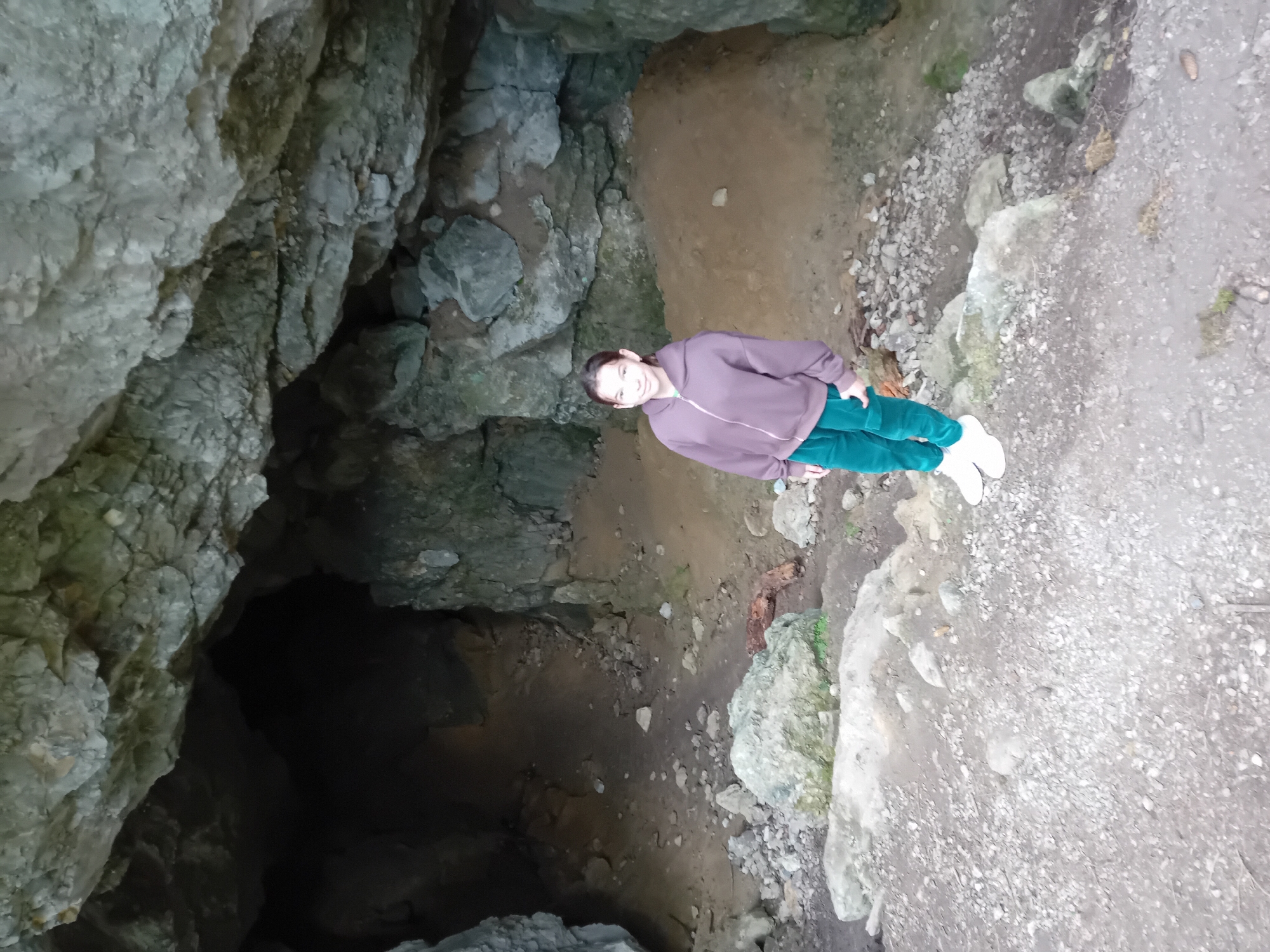 A young girl stands smiling in the mouth of a cave in Zuberec, Slovakia. She is wearing a purple zip-up hoodie and green pants, with white sneakers on her feet. The cave entrance is wide and dark, with rocky formations overhead and on the sides. The ground inside and around the cave is covered in dirt and rocks. The girl is positioned in the center of the frame, facing the viewer, with the cave behind her. The scene appears to be outdoors in a natural setting.