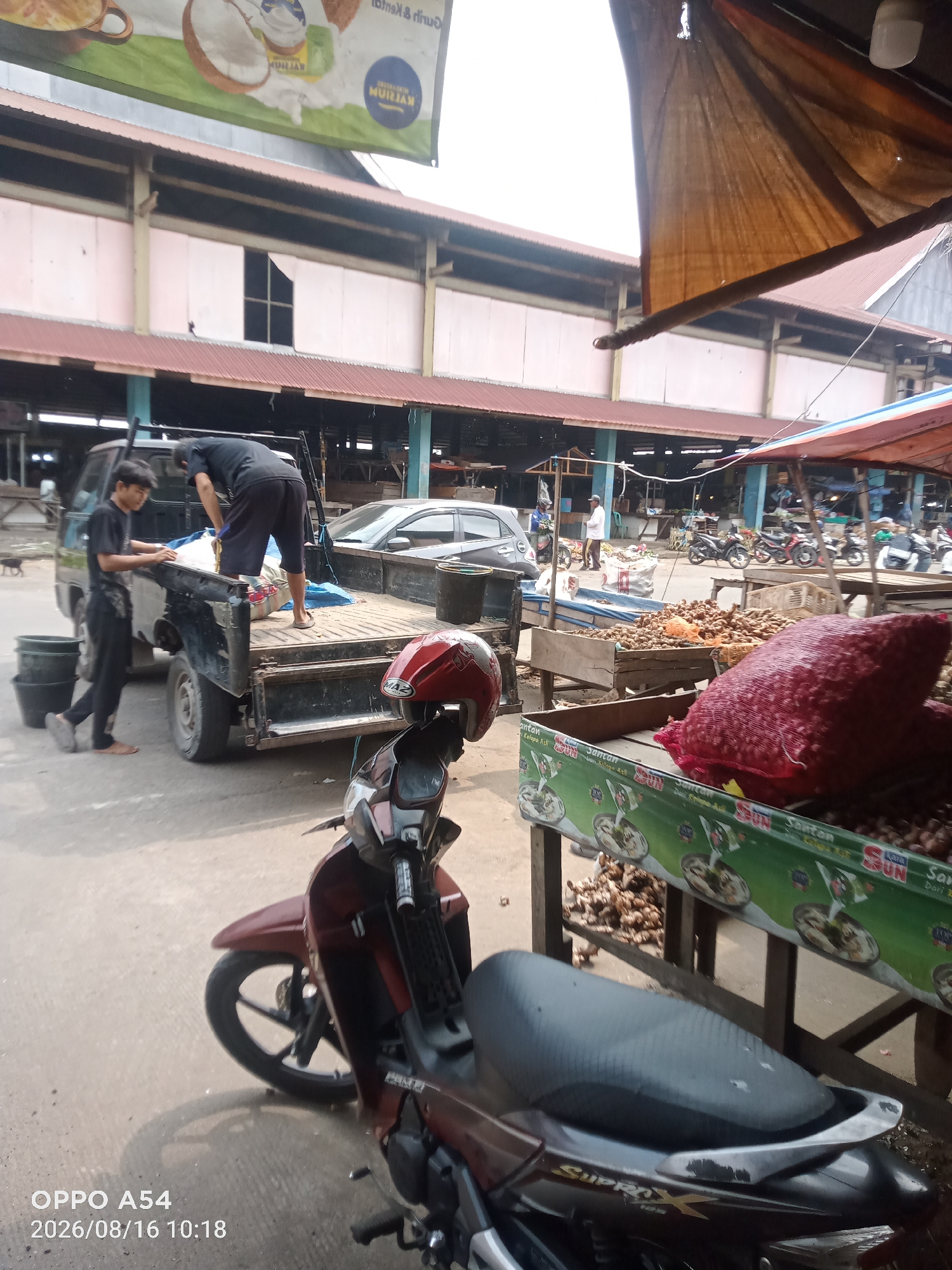 The image shows an outdoor market scene in Jambi, Indonesia. In the foreground, a dark red motorcycle is parked on a paved surface. A person wearing black pants and a black t-shirt is loading items into the bed of a black pickup truck. Another person, wearing black shorts and a black t-shirt, is standing in the truck bed, appearing to adjust or unload goods. Several dark green buckets are on the ground near the truck. A red and white helmet is resting on the back of the motorcycle. In the background, there are market stalls with various goods, including large red bags of produce and piles of root vegetables. A long building with a red roof and light pink walls is visible behind the market stalls. Several other motorcycles are parked in the distance. The overall impression is a bustling marketplace during the daytime.