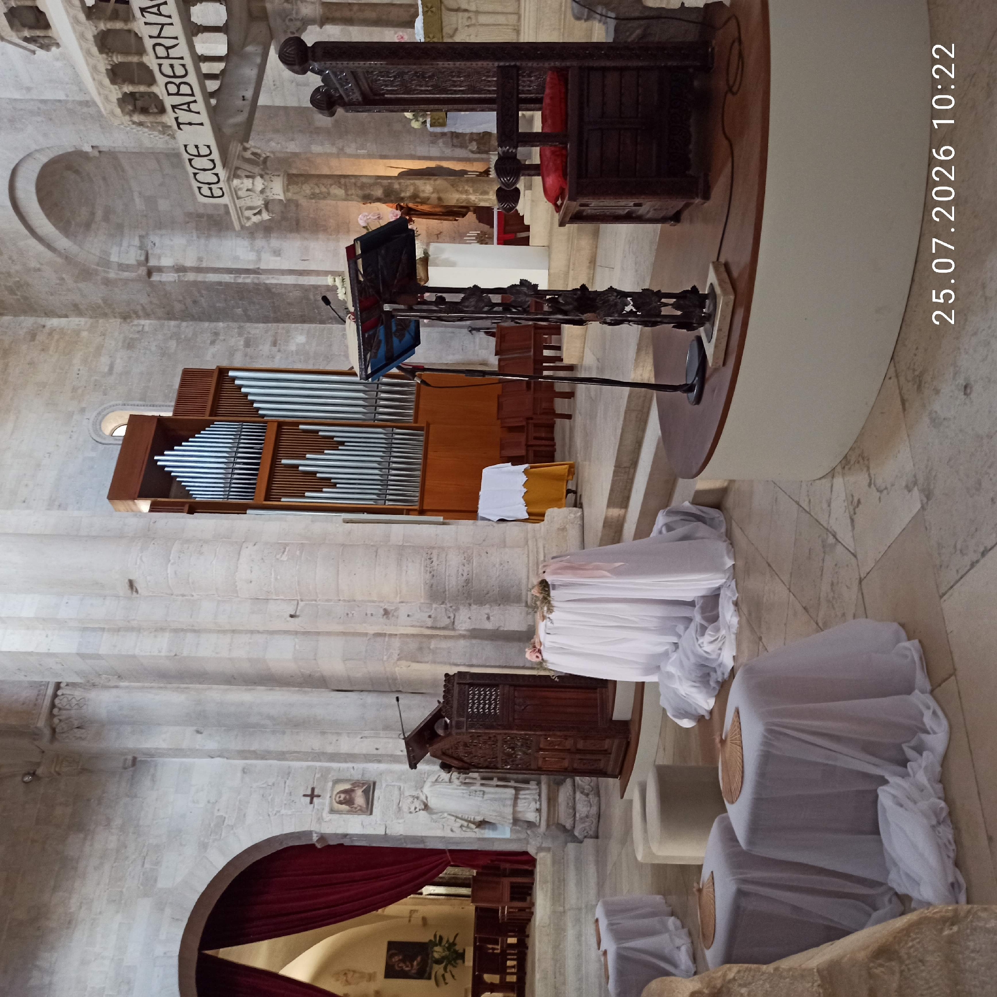 The image shows the interior of the Basilica di Santa Maria Assunta in Ruvo di Puglia, Italy. The scene depicts a section of the church set up for an event, likely a religious ceremony.
In the foreground, to the left, are two square tables draped in white sheer fabric. Each table has a woven fan placed on top. Behind these tables are two cylindrical ottomans. Further to the right, a larger round table is covered in a white cloth and adorned with a pink ribbon and some floral decorations.
Dominating the middle ground is a wooden podium with ornate carvings, positioned on a raised platform. A microphone is attached to the podium. To the right of the podium, a dark wooden ceremonial chair with a red cushion is visible.
In the background, a large pipe organ stands against the stone wall. Above and to the right of the organ, an inscription reads "ECCE TABERNA". The architecture features stone walls, arched doorways, and vaulted ceilings, characteristic of Romanesque style churches.
A statue of a robed figure stands in the background, to the left of the organ, and a framed religious icon is visible on the wall above and to the left of the statue. Red velvet curtains are drawn on the left side of the image, obscuring an archway. Wooden chairs are arranged in the background.
The floor is paved with stone tiles. A date stamp "25.07.2026 10:22" is present in the bottom right corner of the image. No people are visible in the scene.