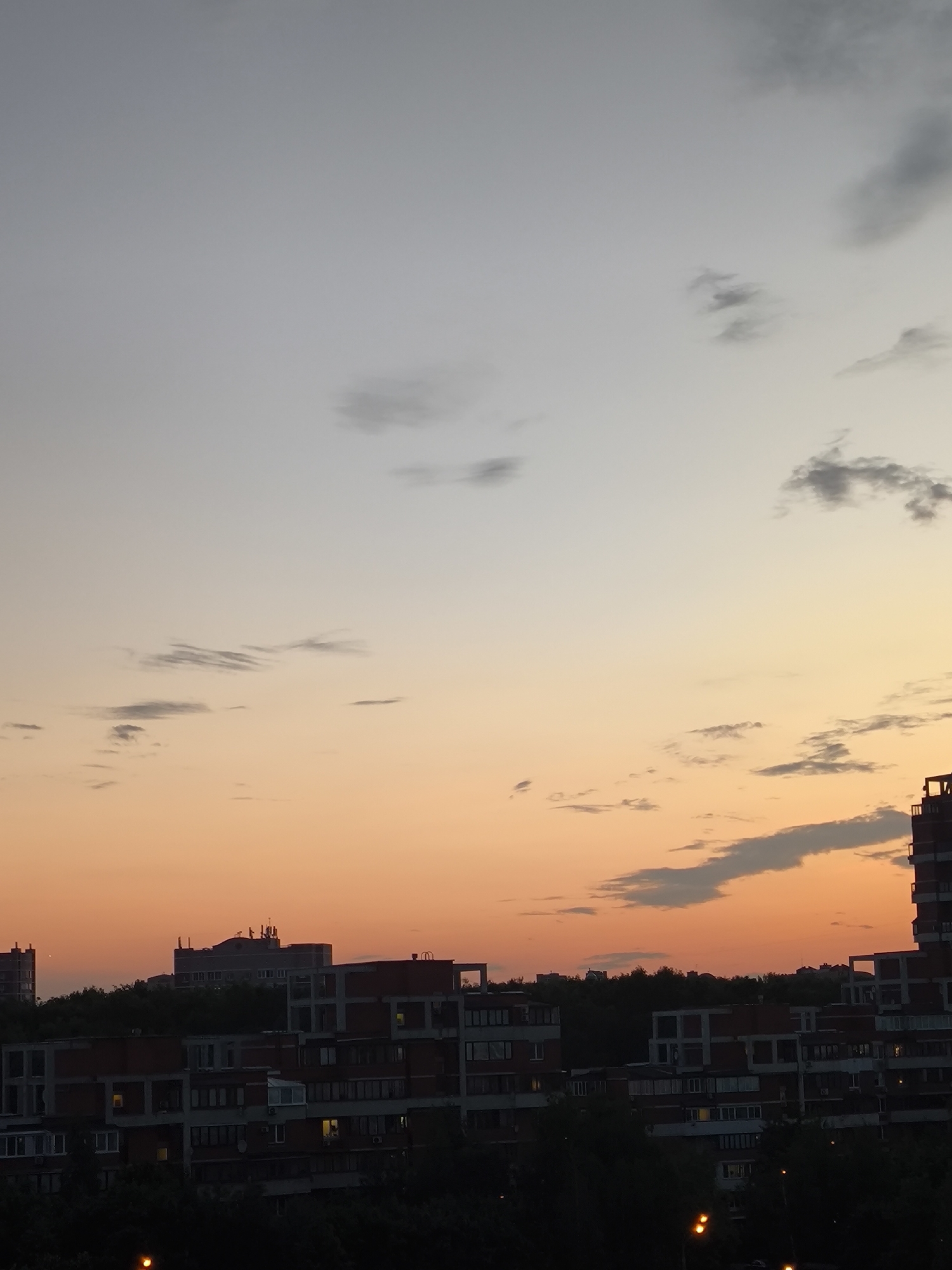 A cityscape at dusk features a gradient sky of light gray at the top transitioning to a vibrant orange near the horizon. Scattered, wispy clouds dot the sky. In the foreground, a dense line of dark trees obscures the base of several buildings. The buildings themselves are predominantly made of concrete and brick, with multiple stories and visible windows, some illuminated. The architecture suggests a residential or apartment complex. The scene is devoid of any visible people or animals. The lighting indicates the setting sun.