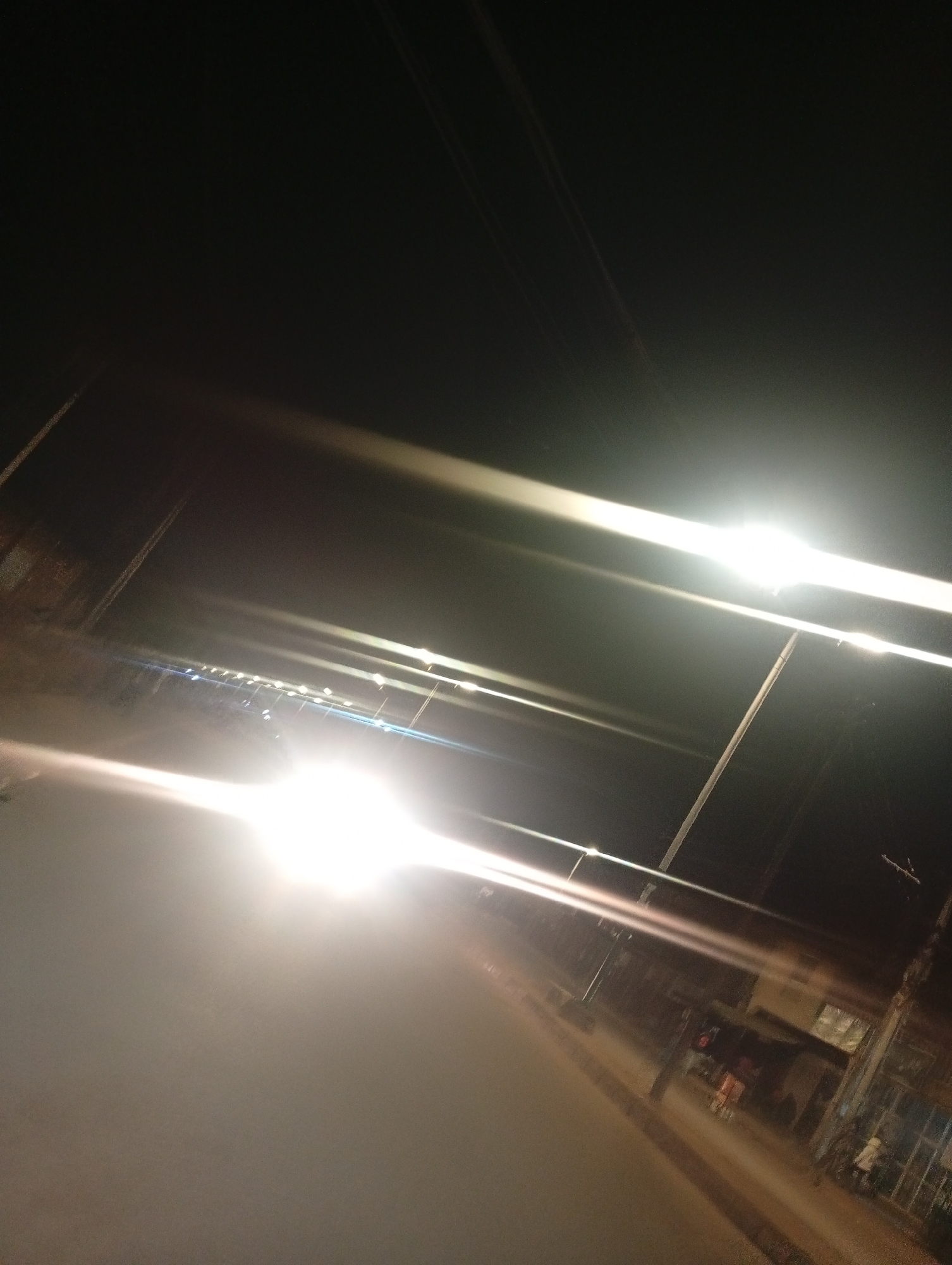 The image depicts a nighttime street scene in Bukuru, Nigeria, illuminated by bright streetlights and possibly vehicle headlights. The perspective is tilted, creating a dynamic and somewhat disorienting view of the environment. The foreground is dominated by a hazy glow from an intense light source, likely a vehicle's headlights, obscuring details of the road surface.
In the middle ground, a line of streetlights stretches into the distance along what appears to be a road. These lights are sharp and elongated due to motion blur or camera angle. Beyond the streetlights, structures that resemble buildings or shopfronts can be faintly discerned, their details softened by the darkness and the bright lights. Some indistinct figures of people are visible near these structures, suggesting activity on the street.
The background is predominantly dark, with the sky being an inky blackness. Thin, faint lines, possibly power lines or wires, are visible against the sky. The overall impression is one of a lively, if somewhat hazy, urban night in Bukuru.