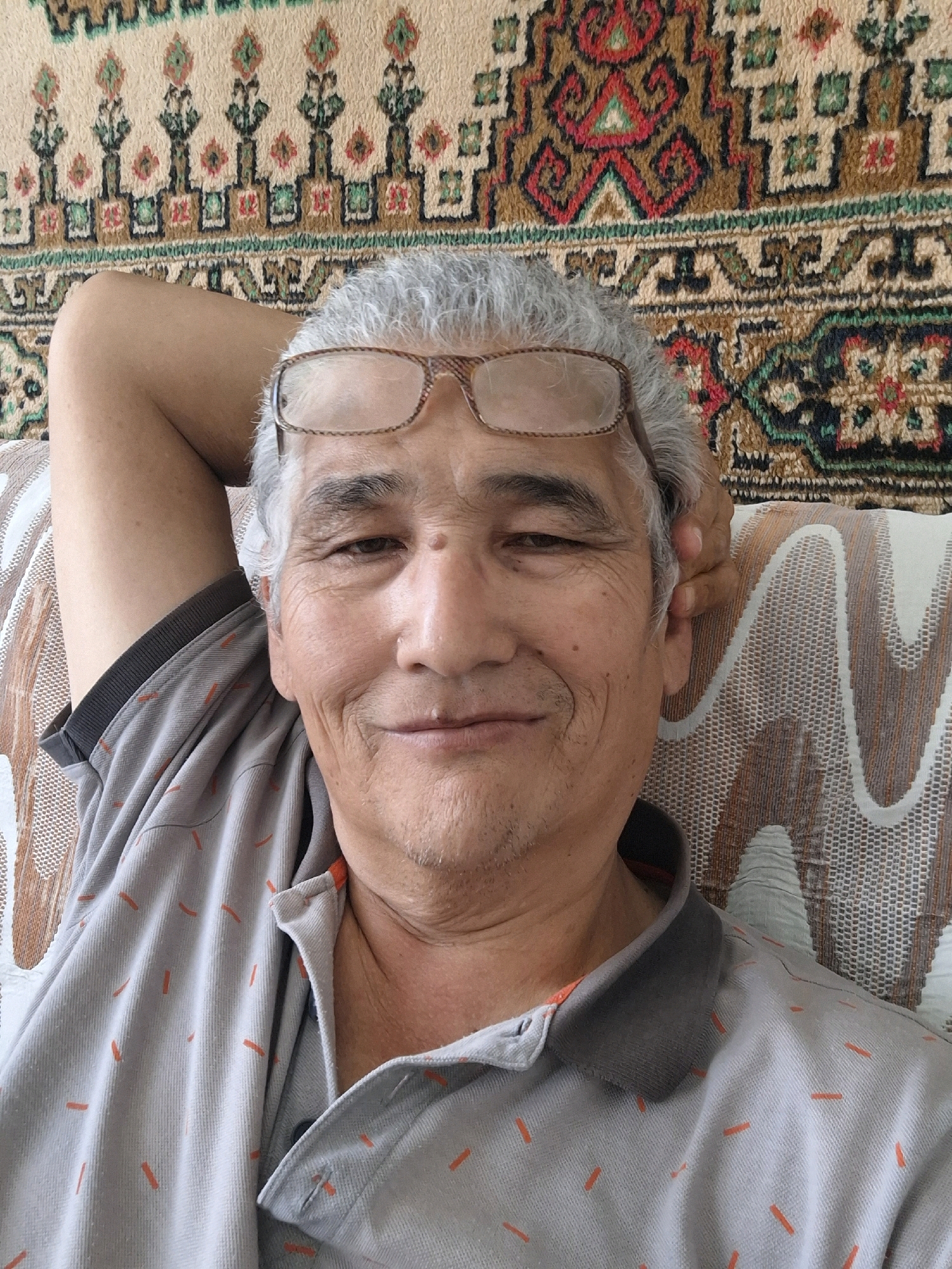 An elderly man with graying hair and glasses is reclining on a couch with his arm behind his head. He is wearing a gray polo shirt with an orange striped pattern. The background features a tapestry with intricate geometric designs and a couch with a patterned upholstery. The scene is indoors, likely in a residential setting in Almaty, Kazakhstan. The man appears relaxed and is looking at the camera.