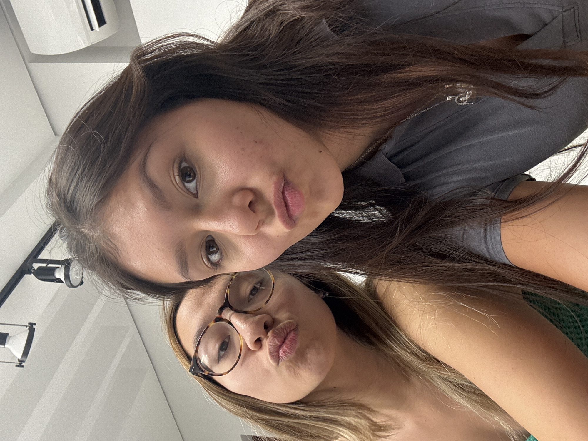 Two young women pose closely together in an indoor setting. The first woman, positioned at the top of the frame, has dark brown hair, fair skin, and is wearing a dark gray shirt. The second woman, located below the first, has lighter brown hair with blonde highlights, wears tortoise-shell framed glasses, and appears to be wearing a green top. Both women are making a kissing face towards the camera. In the background, a white wall with a hanging air conditioner, and an articulated arm holding lights are visible. The specific location within San Andrés Cholula, Mexico, cannot be determined from the given image.