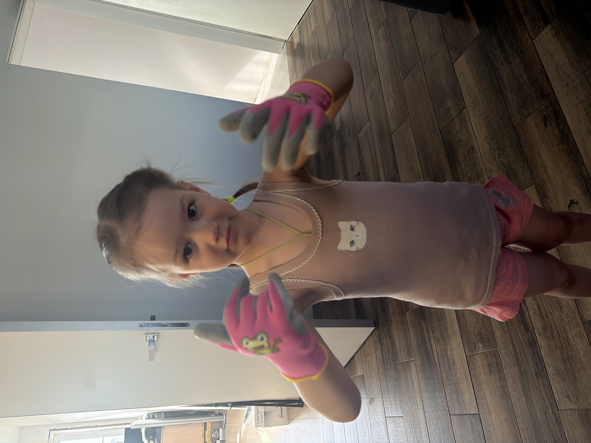 A young girl stands indoors, wearing pink shorts and a tan tank top with a cat face graphic. She has light hair pulled back in a ponytail with green bands. The girl is wearing pink and grey gardening gloves and holding her hands up in front of her. The background consists of a closed white door, part of a grey wall, and wood flooring. There is a glimpse of what appears to be kitchen cabinets through a doorway. The image was likely taken inside a residence in Chaikovsky, Russia.