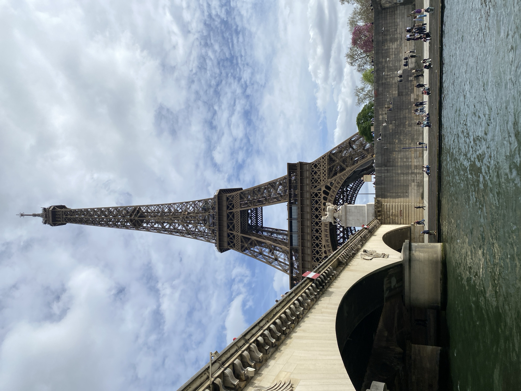 The image shows the Eiffel Tower in Paris, France, as viewed from a low angle near or on a bridge over the Seine River. The tower occupies much of the left side of the frame, ascending towards the cloudy, partially blue sky. The stone bridge features ornate details, including decorative balustrades and archways over the river. Along the riverbank, visible on the right, are stairs where people are sitting and standing, interacting and enjoying the view. The river, with a greenish hue, fills the lower right portion of the frame. The overall composition emphasizes the height and architectural detail of the Eiffel Tower in relation to the surrounding urban landscape.