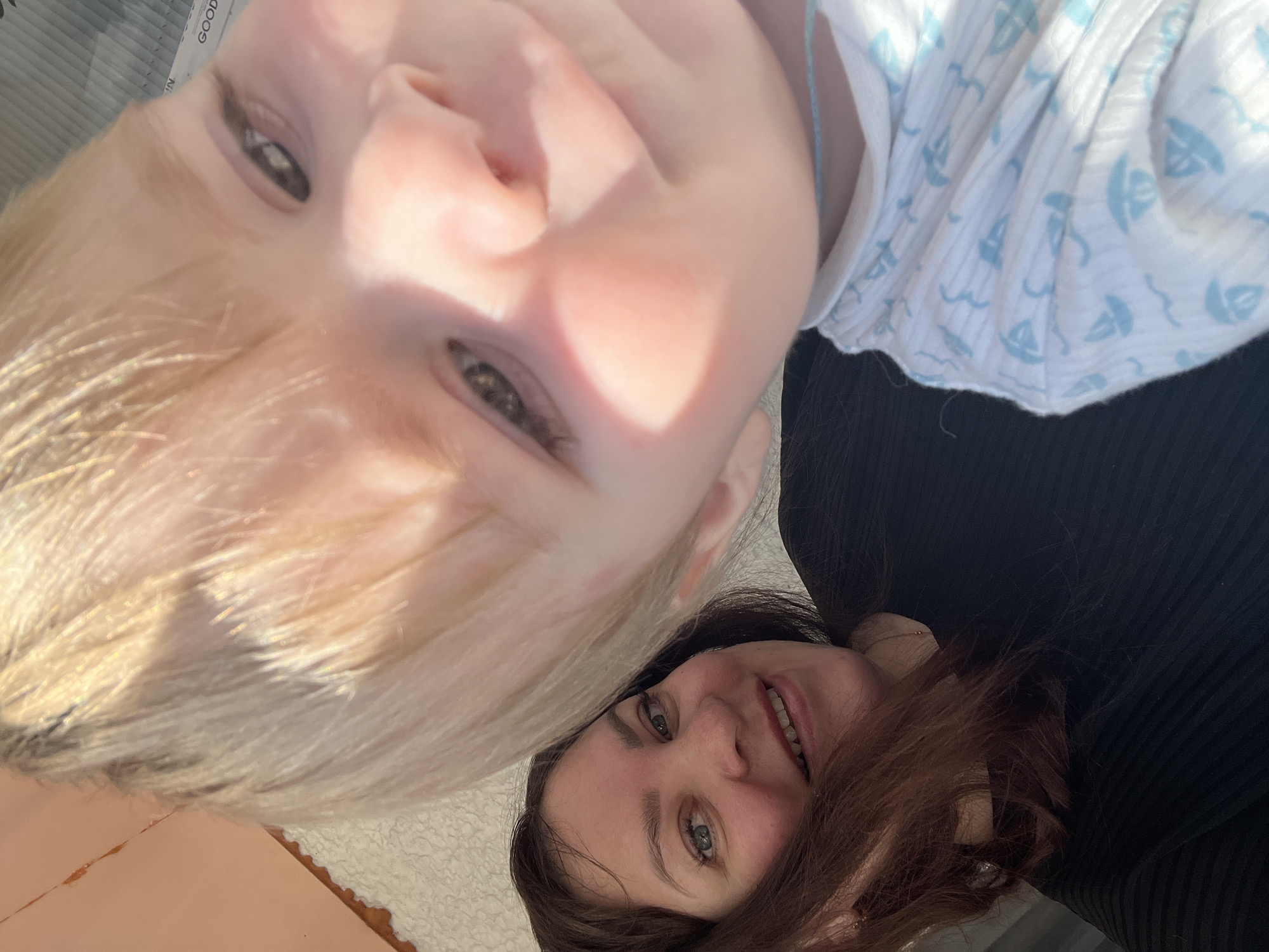 The image shows a close-up selfie of two individuals, an adult and a child, likely outdoors. The adult, positioned to the left and slightly behind the child, has long, brown hair and is wearing a black garment. They are smiling and looking towards the camera. The child, in the foreground and to the right, has blonde hair and is also looking towards the camera, with a partially visible face. The child is wearing a white garment with a light blue pattern of sailboats. Sunlight is casting shadows and highlights on their faces, suggesting a bright, outdoor environment. There is no discernible urban infrastructure or specific landmark that would indicate a precise location within Noviy, Russia.