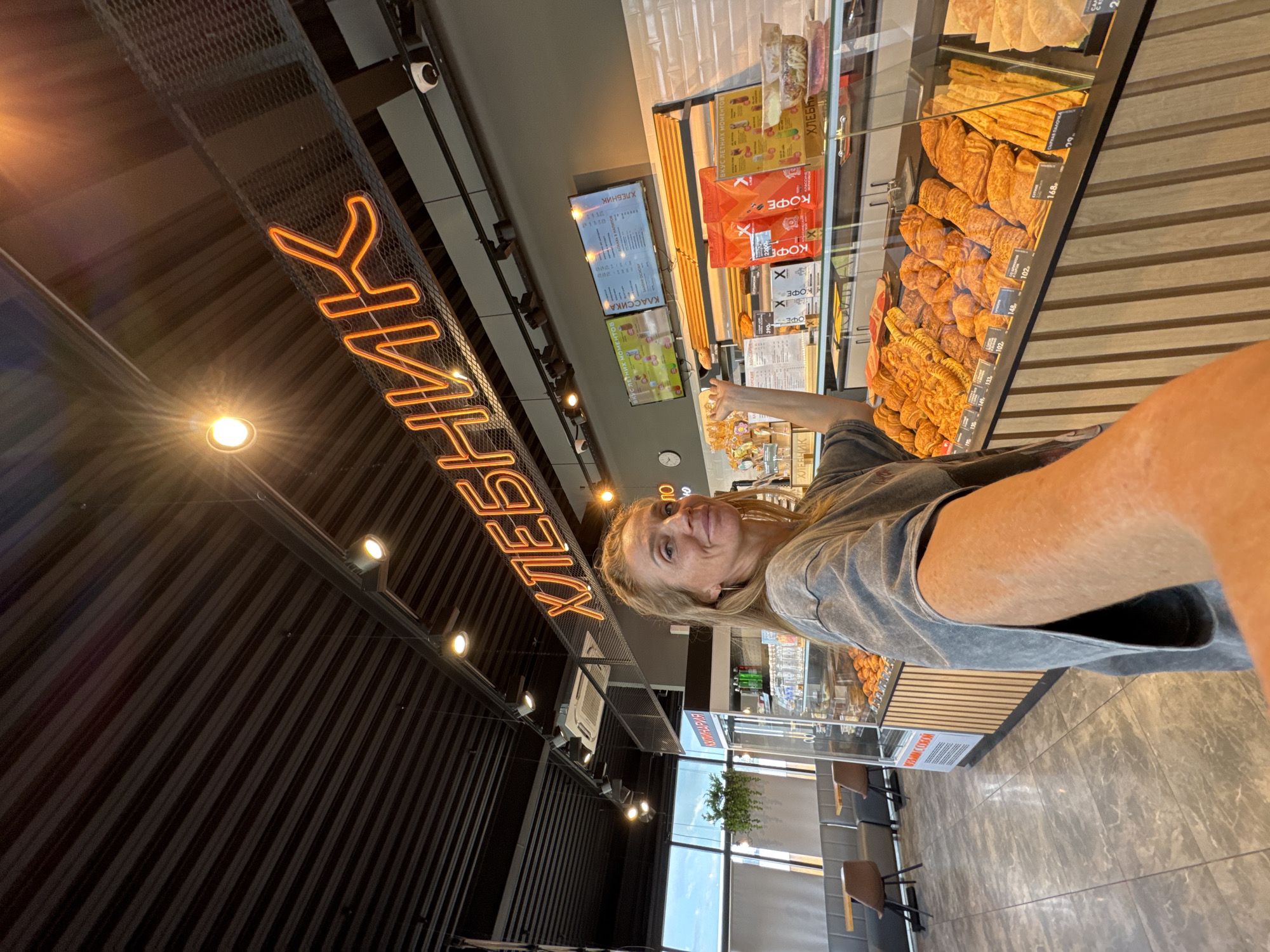 This media file captures a selfie taken inside a modern bakery or cafe in Peski, Russia. The primary subject is a light-haired person, likely female, smiling faintly at the camera while pointing with their right hand towards a glass display case brimming with a variety of golden-brown baked goods, including croissants and various rolls.
The setting is a well-lit, contemporary interior with a dark, ribbed wall texture. A prominent orange neon sign on the ceiling spells out "ХЛЕБНИК" (Khlebnik), which means "baker." Below it, multiple digital screens display menus and advertisements, featuring text like "КУЛИНАРИЯ" (Culinary), "КЛАССИКА" (Classics), and "ВКУС ЛЕТНИХ МОМЕНТОВ" (Taste of Summer Moments). Various packages of "КОФЕ" (Coffee) are also stacked near the display. Price tags, such as "102" and "168," are visible next to the pastries. The floor is a light grey, mottled tile, and through large windows in the background, a seating area with dark chairs and tables is visible, suggesting natural daylight. The overall ambiance is modern and inviting, indicating a pleasant experience for the person taking the selfie. A security camera is also visible on the ceiling.
