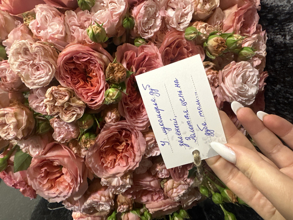 A bouquet of roses in shades of pink and peach fills the frame. A small, rectangular tag rests among the flowers, bearing handwritten Cyrillic text in blue ink. A hand with long, oval, white-painted nails holds the tag in place. The background is dark, possibly a table or surface, emphasizing the colors and texture of the roses.
