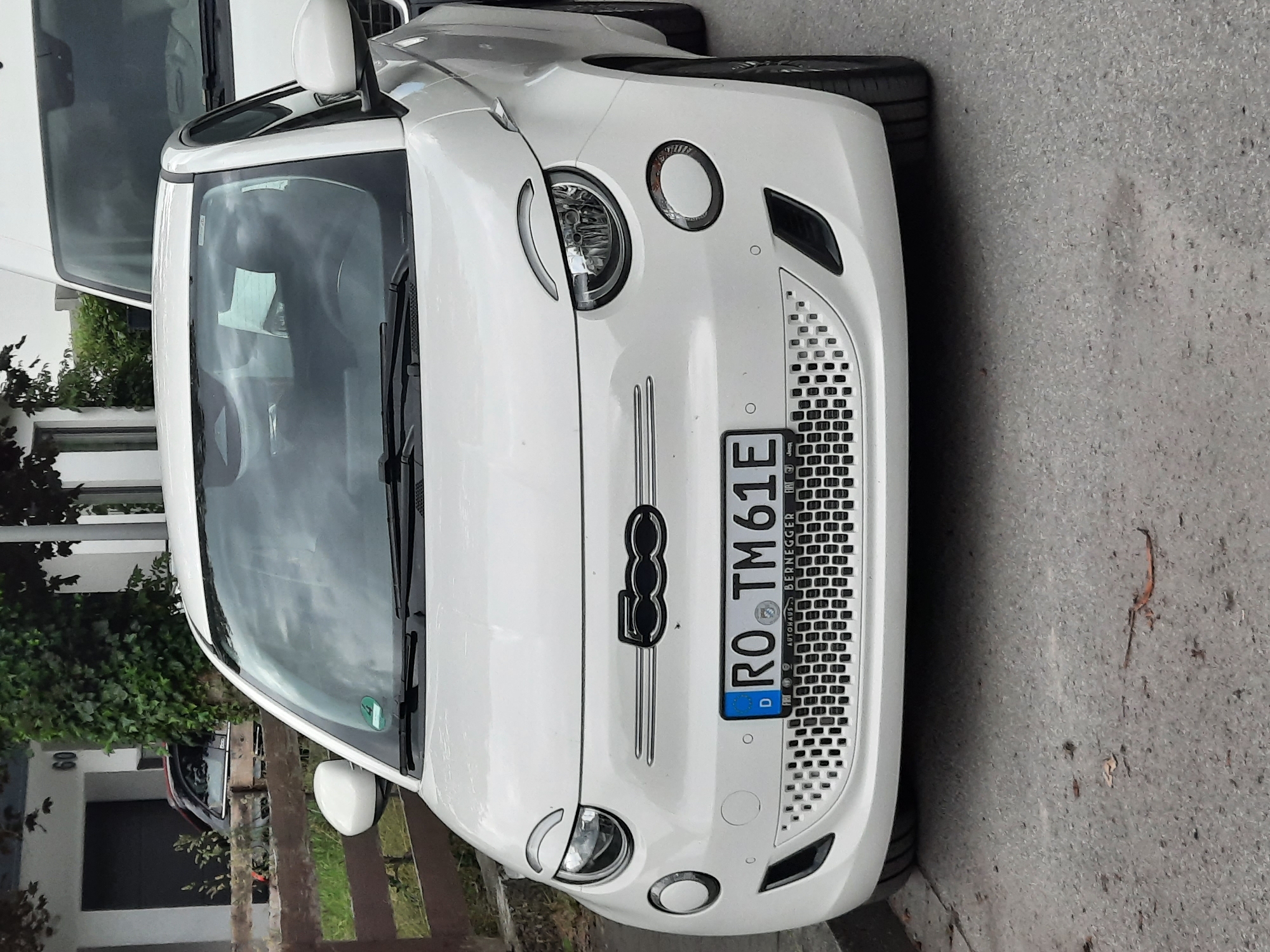 A white Fiat 500 electric car is parked on a street in Rosenheim, Germany. The car is facing forward, and its license plate reads "RO TM 61E". The car has a black grill with a honeycomb pattern and round headlights. In the background, there is a building with the number 60 on it, and a wooden fence. To the right of the Fiat, a white van is partially visible. The street appears to be paved with asphalt, and there are some fallen leaves on the ground. The overall impression is that of a typical urban street scene.