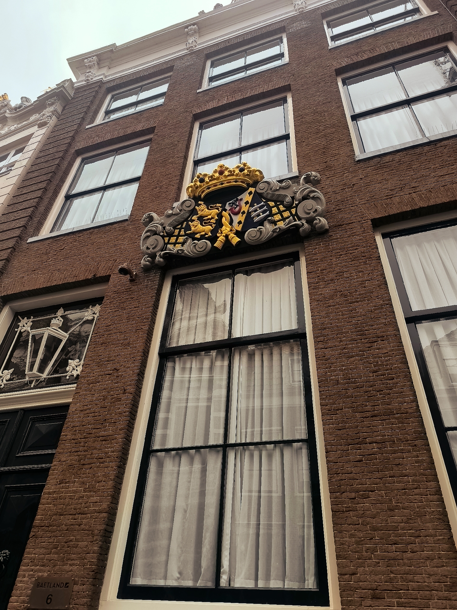 The image shows the facade of a brick building with several windows. Above one of the windows is a coat of arms with a crown, lions, and shields. The building appears to be in a historic district. A sign at the bottom left of the image reads "BAETLAND 6". This is likely an address in Hoorn, Netherlands. The sky is overcast.