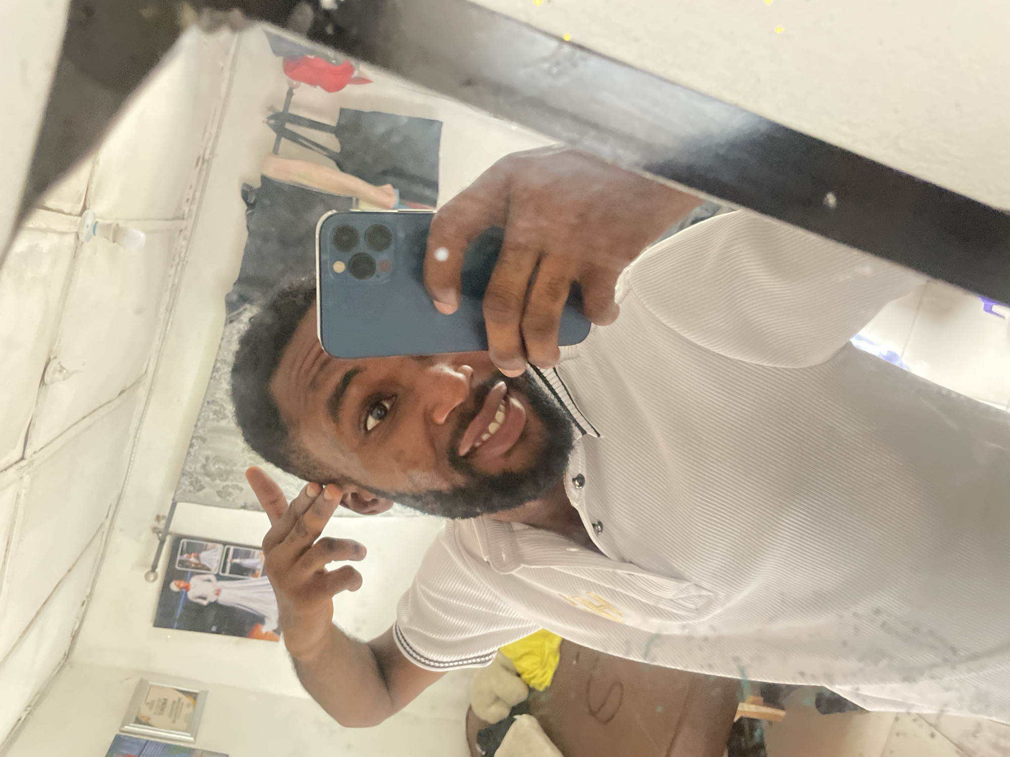 A person is taking a selfie in a mirror with a blue smartphone. The person has dark skin, short dark hair, and a beard. They are wearing a white shirt with a striped pattern. The background appears to be a room with white walls. There are various items visible in the reflection, including a red object, clothes, and framed pictures. The location is not discernible as Lokoja, Nigeria, based solely on the information in the image.
