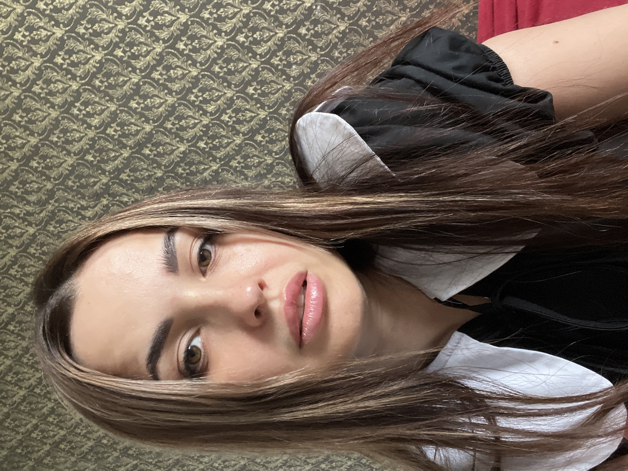 The image shows a person with long, light brown hair looking directly at the camera. They have brown eyes and are wearing a black top with a white collar. The background consists of a wall with a patterned design, and a red surface is partially visible behind the person. The location and city are unknown.