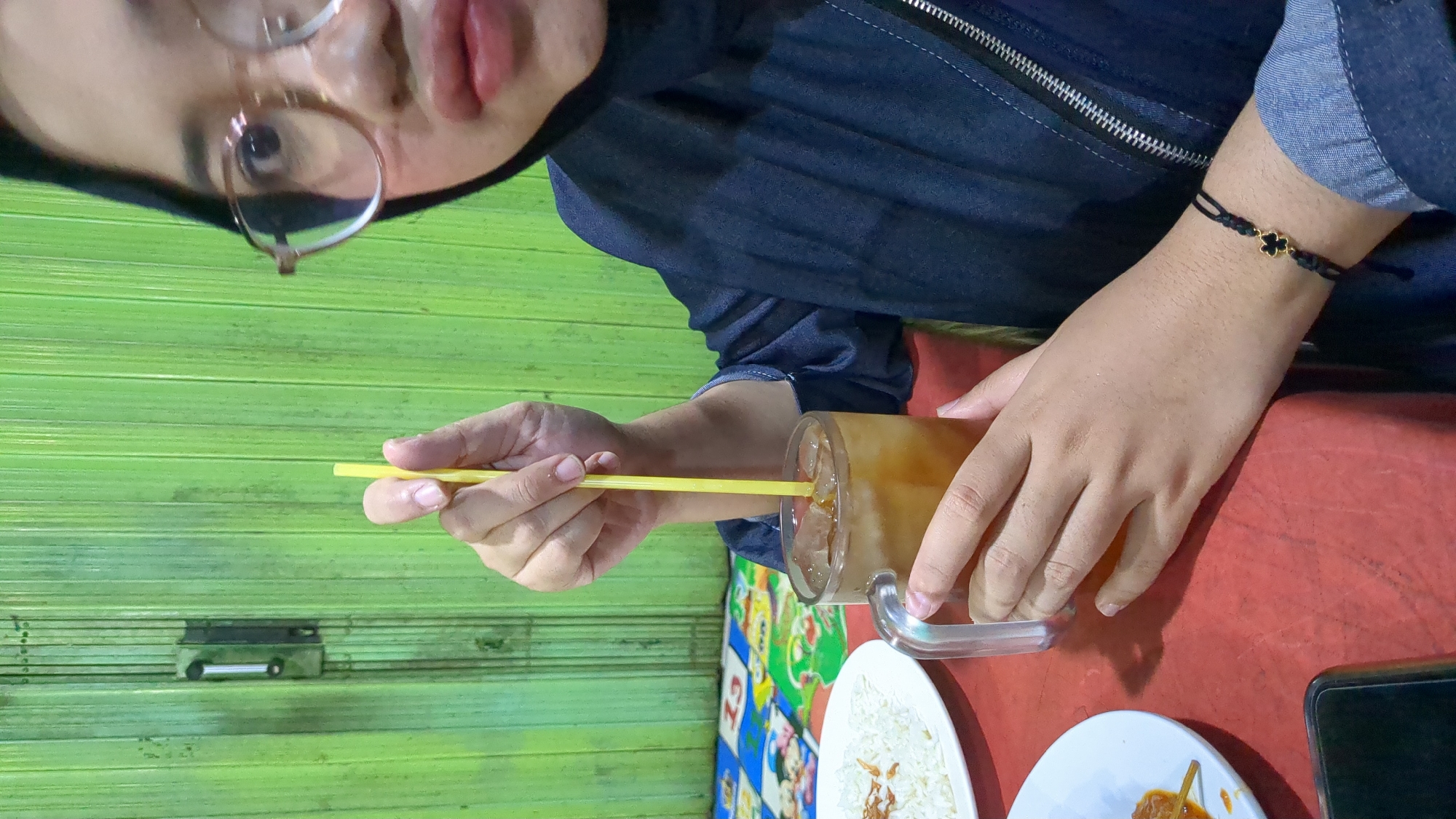 A close-up, medium shot shows a person, wearing a hijab and glasses, sitting at a red table. The person is holding a glass of iced tea with a yellow straw. To the left, a plate of white rice is visible. The background features a green metal accordion door. The scene appears to be a casual dining setting in Purwakarta, Indonesia.