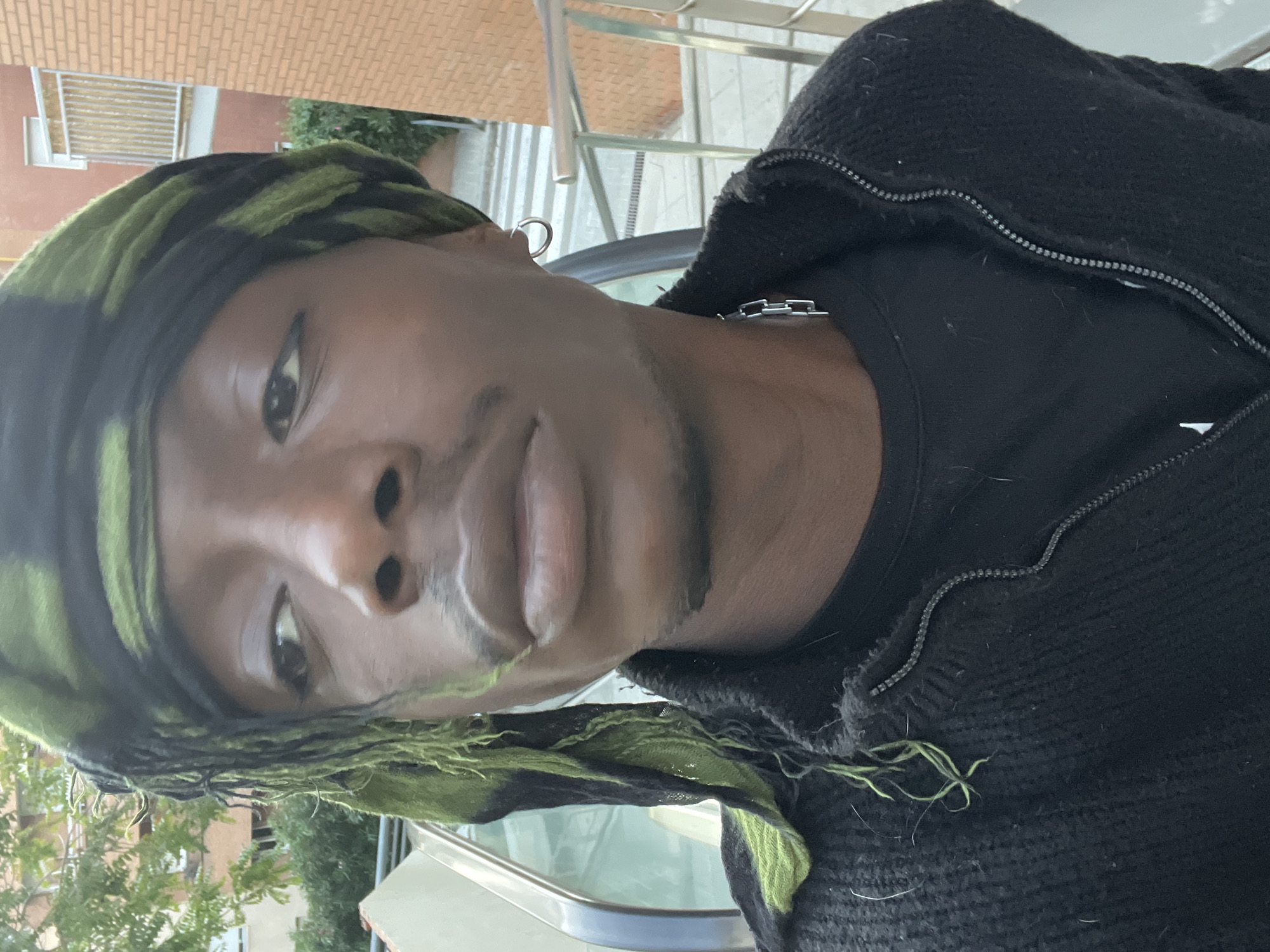 A person with brown skin, wearing a black t-shirt under a black zip-up sweater, and a green and black headscarf is the primary subject of this photo. A silver hoop earring is visible on their left ear and a silver chain necklace on their neck. The background reveals an urban environment, featuring a light-colored brick building and greenery. A metal railing and the top of a white vehicle are also visible, along with an additional building in the background. There is not enough information to conclude this photo was taken in Barcelona, Spain.