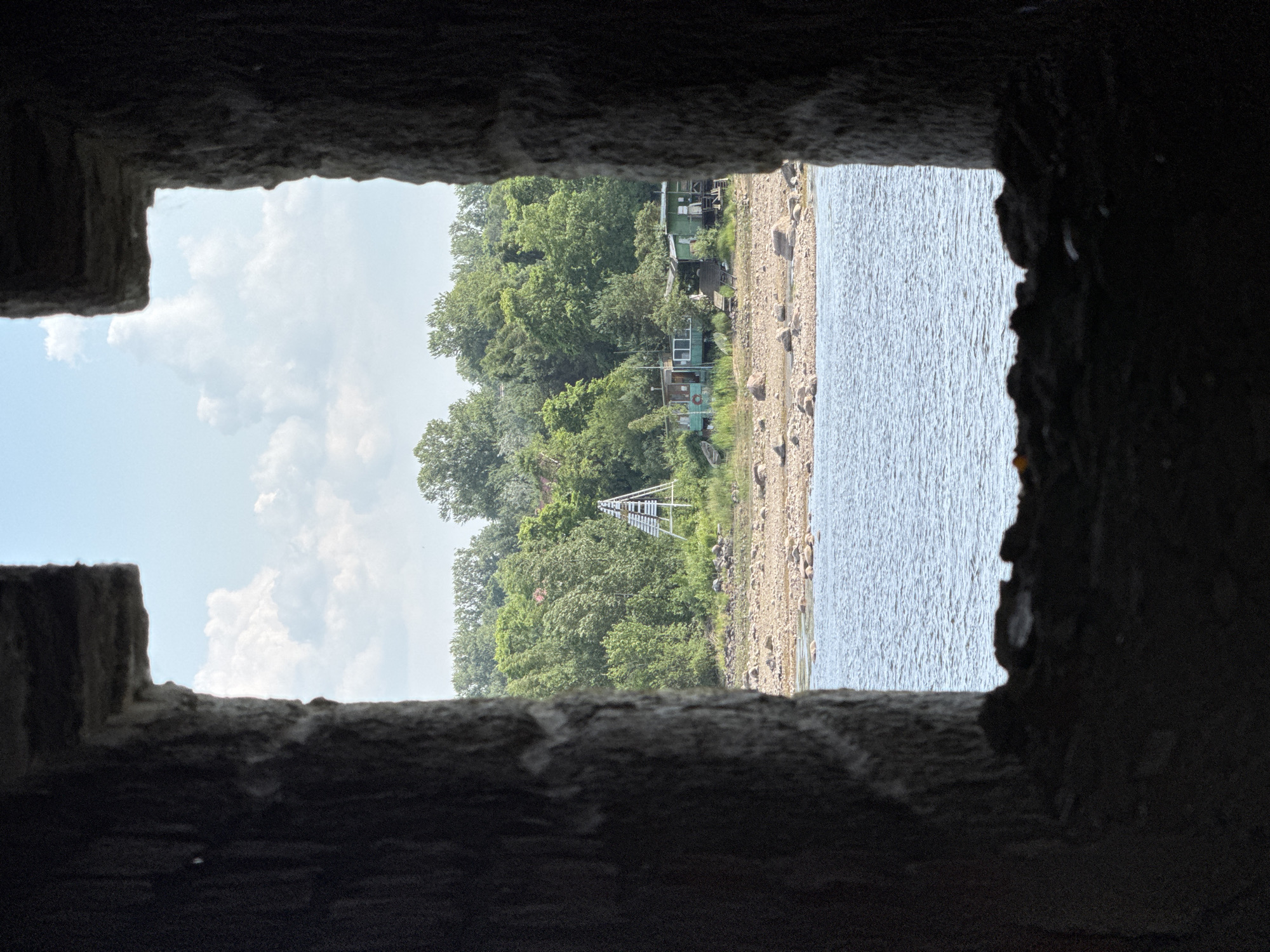 The image shows a view through a rough stone opening, looking out onto a bright, cloudy sky and a body of water. The opening frames a landscape with a tree-lined shore, a rocky beach, and several small buildings nestled amongst the trees. A white, pyramid-like structure, possibly a lookout or observation post, is visible in the distance. The water is calm with visible ripples, suggesting a gentle breeze. The overall impression is one of a quiet, natural setting, possibly a lakeside or riverbank. The time of day appears to be daytime, with bright daylight illuminating the scene. The stonework of the opening is textured and appears aged, adding a sense of history or permanence to the view.