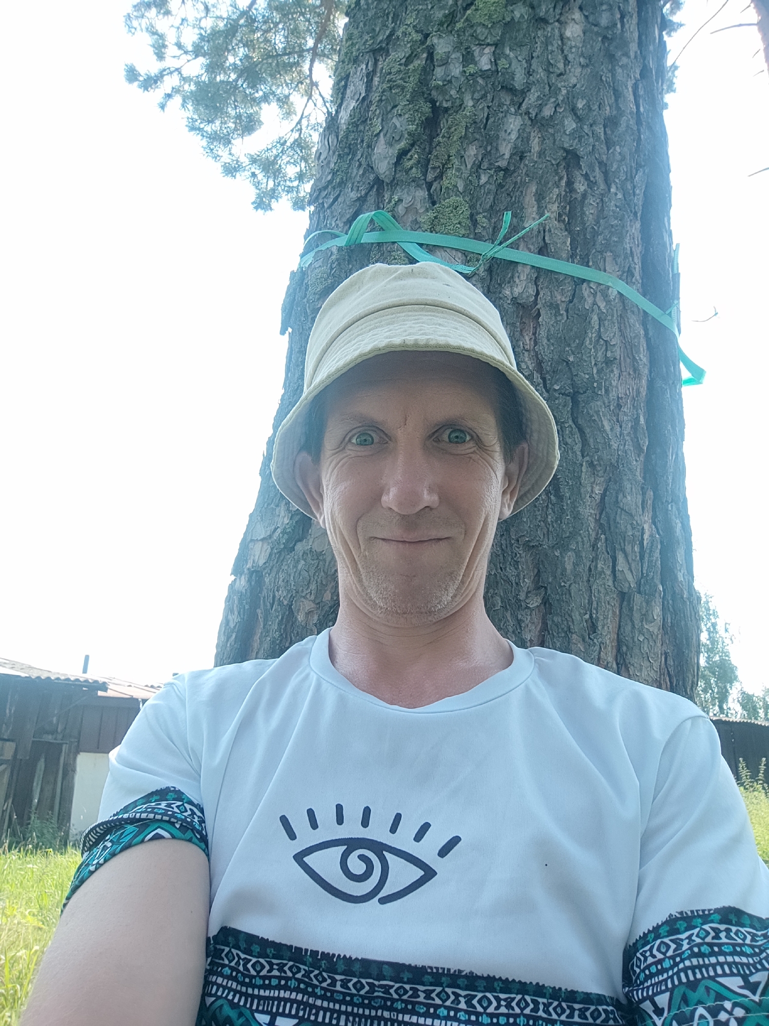 A man wearing a light-colored bucket hat and a white t-shirt with a black eye design and patterned sleeves is leaning against a large tree. A green strap is tied around the tree. The man is smiling and looking at the camera. In the background, there is grass, a wooden structure that appears to be a fence or shed, and trees. The sky is bright and appears overcast or washed out. The scene is outdoors, likely in a rural or semi-rural area, possibly the city of Sokolovo, Russia, given the context.