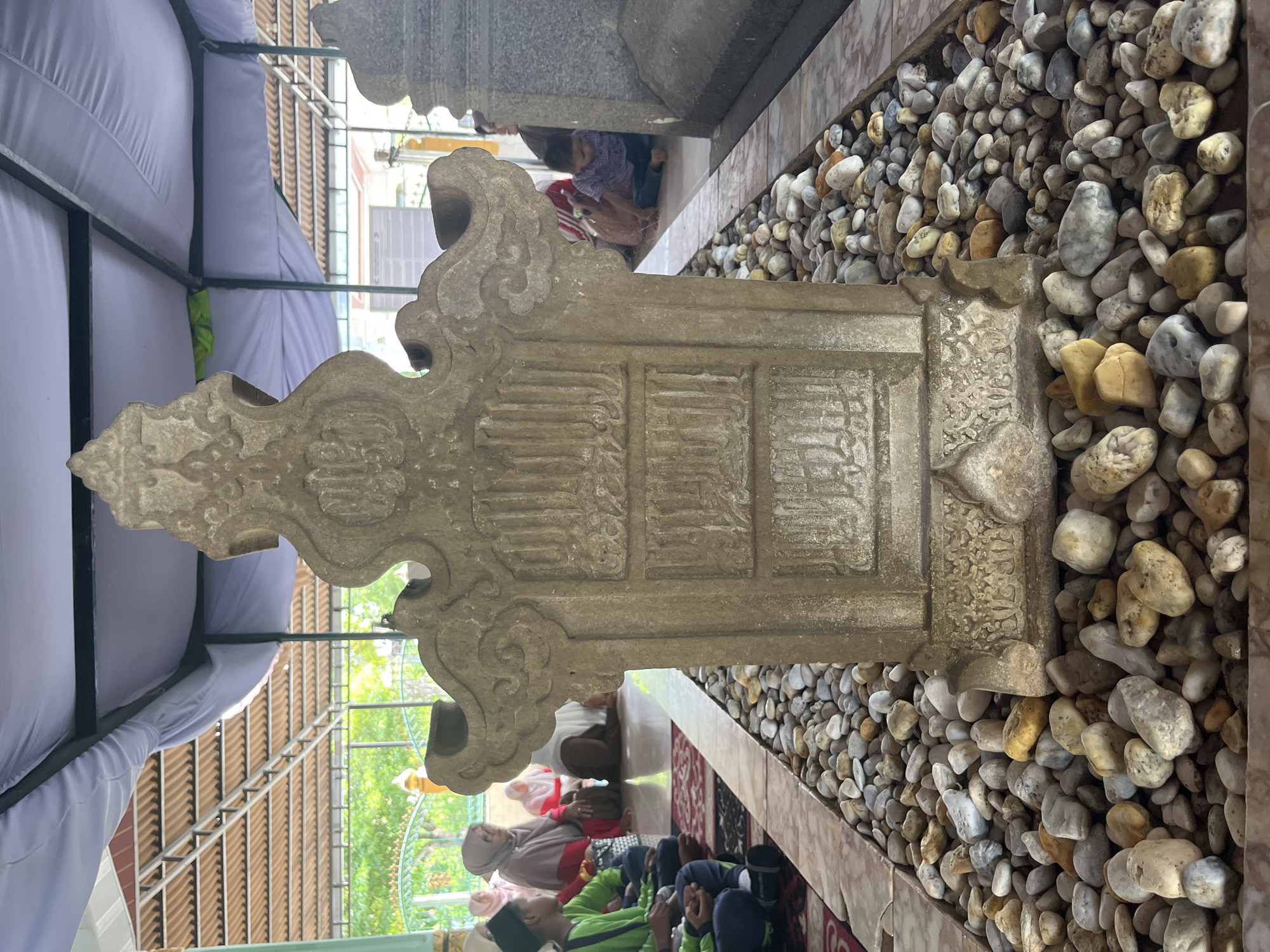 The image depicts a tombstone in the city of Beuringen, Indonesia. The tombstone, carved from light-colored stone, is intricately designed with Arabic calligraphy. It is surrounded by a bed of smooth, rounded pebbles of various colors, including grey, white, and yellow. The tombstone is situated outdoors, beneath a canopy structure supported by metal beams and covered with a light purple fabric. In the background, several individuals, including men, women, and children, are seated on the floor, some wearing traditional Islamic attire. The ground appears to be tiled, with a red carpet visible in the foreground. The overall environment suggests a sacred or commemorative site, possibly a cemetery or a shrine.