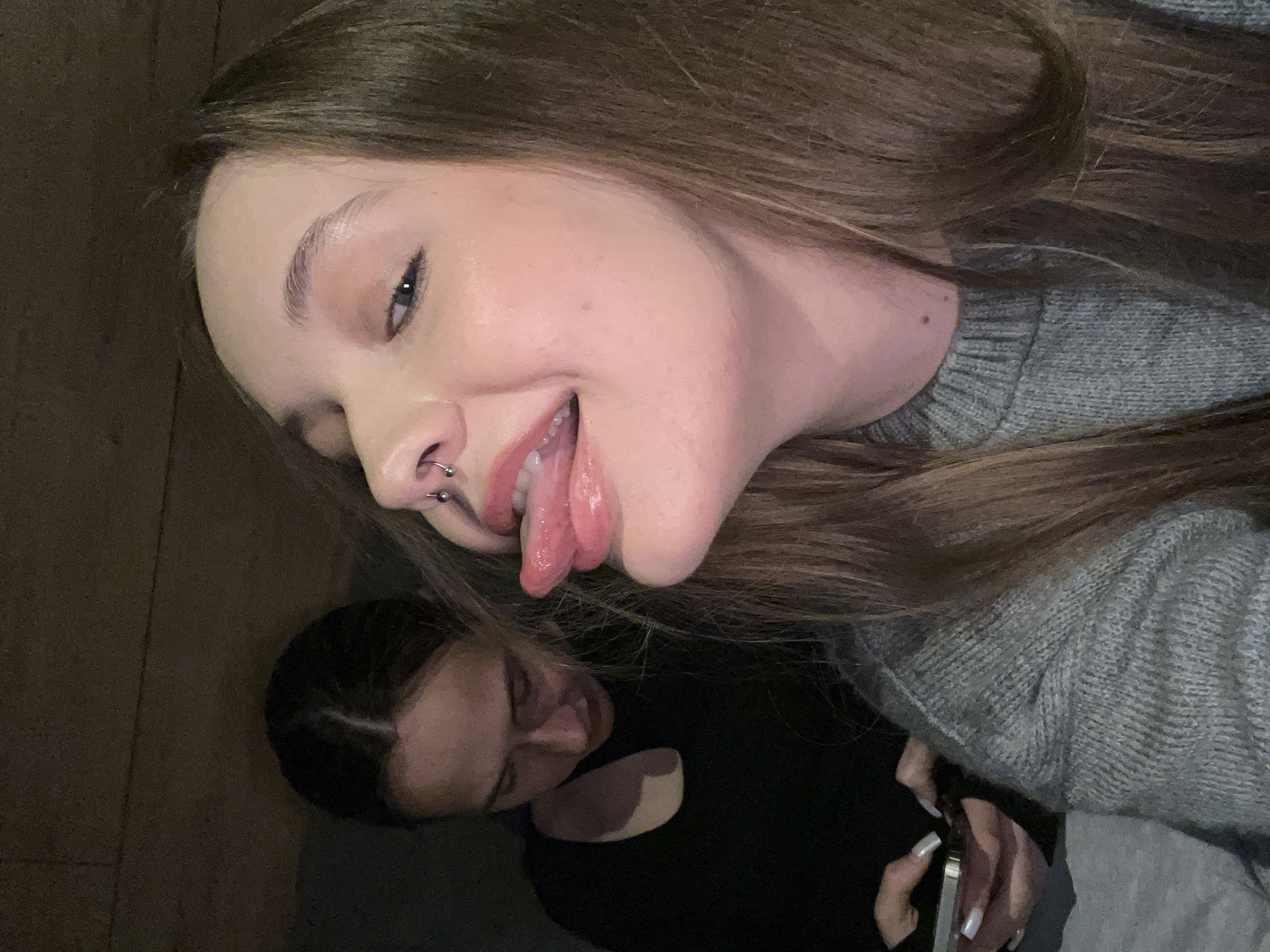 Two women are captured in an indoor setting. The primary subject, positioned in the foreground, is a woman with light brown hair, a nose piercing, and wearing a gray sweater. She is sticking out her tongue. Slightly blurred in the background is another woman with dark hair, wearing a black top, and holding a smartphone. The location is in the city of Unknown, Unknown.