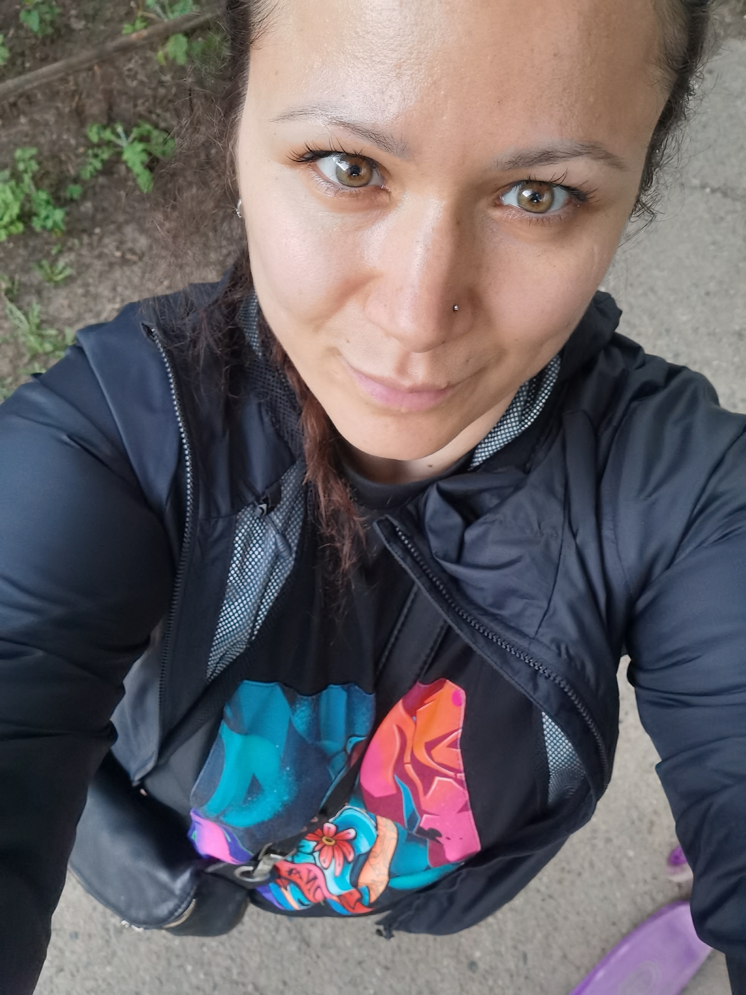 A woman with brown hair pulled back into a bun, brown eyes, and a nose piercing is taking a selfie. She is wearing a black jacket over a black t-shirt with a colorful graphic design. A black bag hangs from her shoulder. The woman is standing on a concrete surface. In the background, there is some green vegetation. A portion of a purple object, potentially a skateboard, is visible in the lower right corner. Given that the request specifically states the city of Chaikovsky, Russia, it is possible that this photo was taken there, though this is not evident from the image itself.