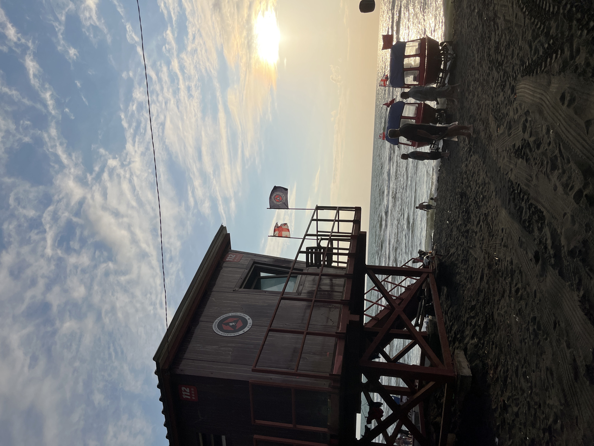 The image captures a view of a black sand beach in Ureki, Georgia, with a lifeguard station in the foreground. The lifeguard station is a wooden structure on stilts, painted red and brown, with a balcony and a chair visible. Two flags are flying from poles attached to the balcony of the lifeguard station. One flag is white with a red cross and red emblems in the corners, while the other is dark blue with a circular emblem. The sea stretches out to the horizon, with gentle waves lapping at the shore. The sun is setting, casting a warm glow across the sky and sea, with visible sun flares.
In the midground, several people are gathered on the beach. Some are standing near two small, red boats on trailers, which appear to be motorboats or similar watercraft. Other individuals are seen walking or standing closer to the water. One person is in the shallow water. The beach itself is composed of fine, dark sand, with tire tracks visible from vehicles. The sky is a mix of blue with scattered white clouds, and the overall atmosphere suggests a late afternoon or early evening scene.