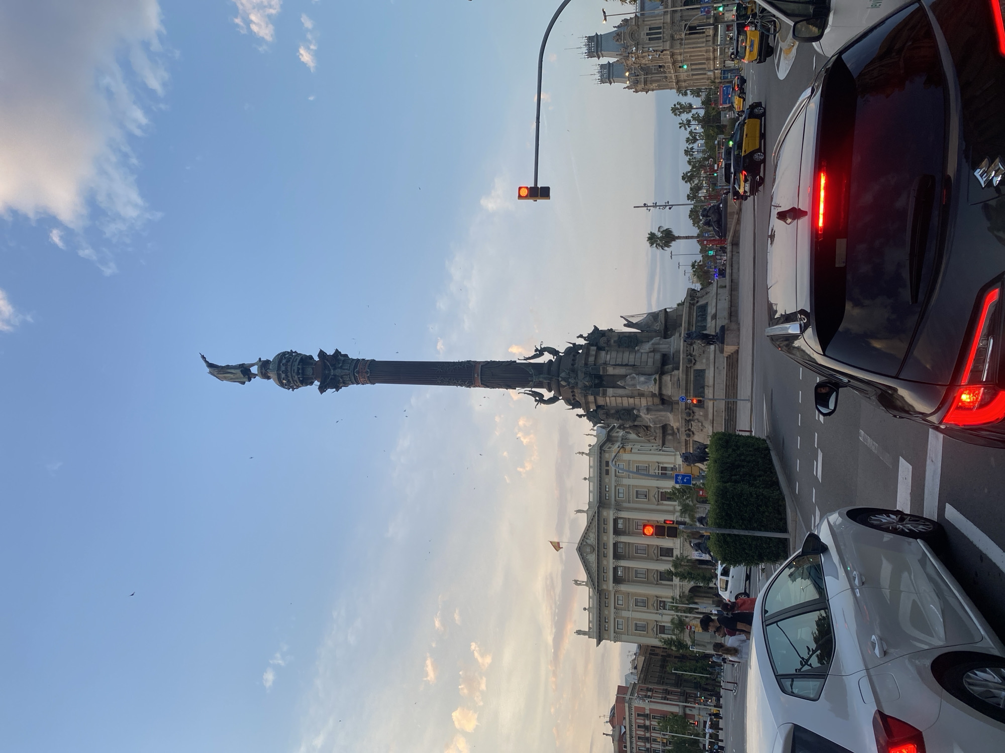 The image depicts a street-level view of the Columbus Monument in Barcelona, Spain, at dusk. A tall, ornate column topped with a statue of Christopher Columbus stands prominently in the background. Several vehicles are stopped at a traffic light, with the red light illuminated. In the foreground, the rear of a dark-colored car is visible, along with the side of a white car. People can be seen walking on the sidewalk in the mid-ground, and a lush green hedge lines the street. The sky is a soft blue with scattered clouds, and a few birds are flying overhead. The architectural style of the buildings surrounding the monument suggests a historical European city.
