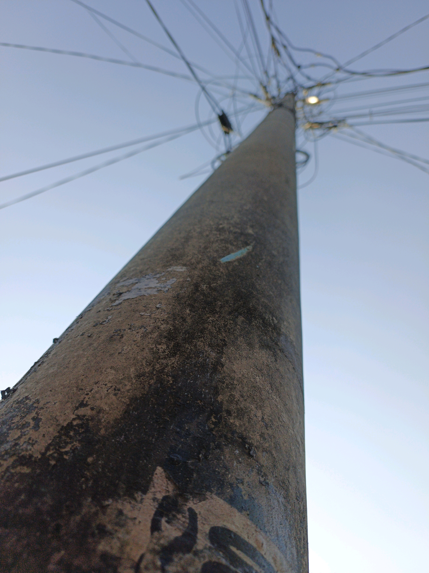 This is a low-angle, close-up shot of a weathered concrete utility pole in Ketegan, Indonesia. The pole is textured with patches of peeling paint and dark stains, suggesting age and exposure to the elements. A dense network of black electrical wires and cables emanates from the top of the pole, fanning out against a clear, pale blue sky. One light bulb is visible near the top, faintly illuminated. The image focuses on the texture and scale of the pole, emphasizing its height and the complex web of wires it supports.
